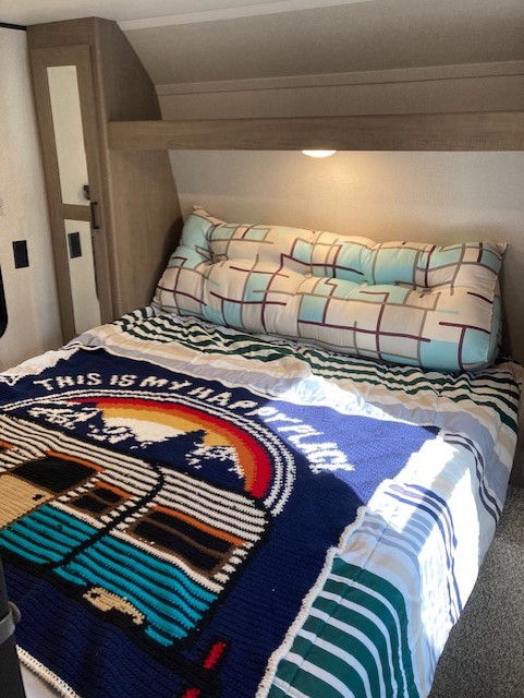 Cozy RV rental bedroom with "Happy Place" blanket, bolster pillow, and overhead storage for comfortable stays.. Keystone RV Hideout 2021