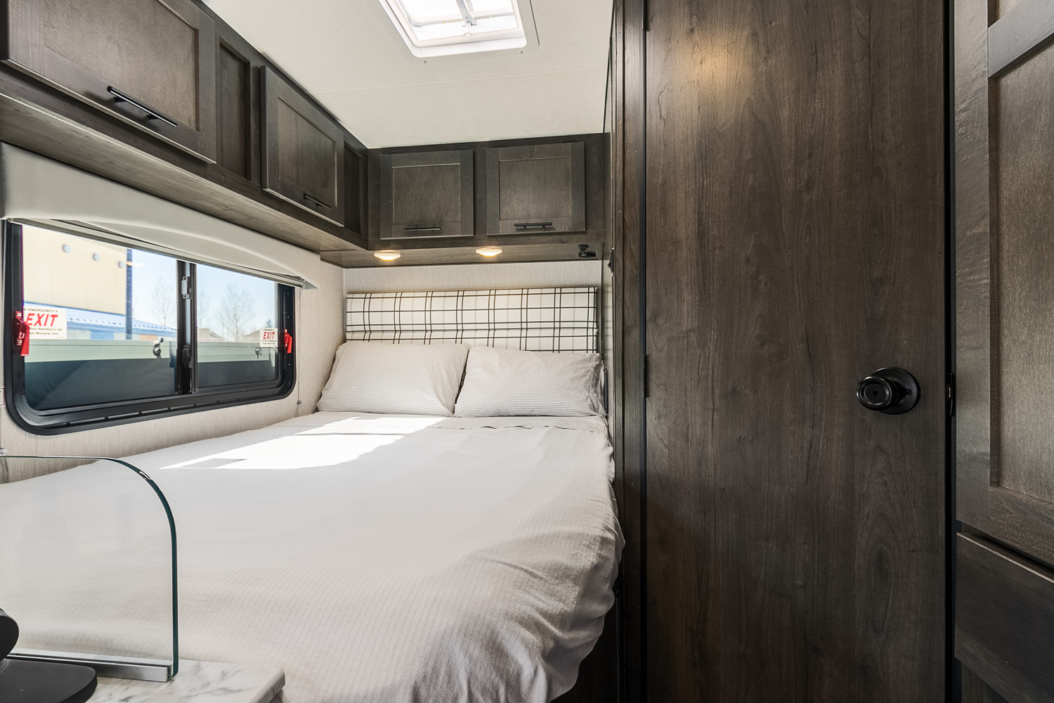 Cozy RV rental bedroom with a comfortable bed, plaid headboard, dark overhead cabinets, and a large window.. Other Other 2023
