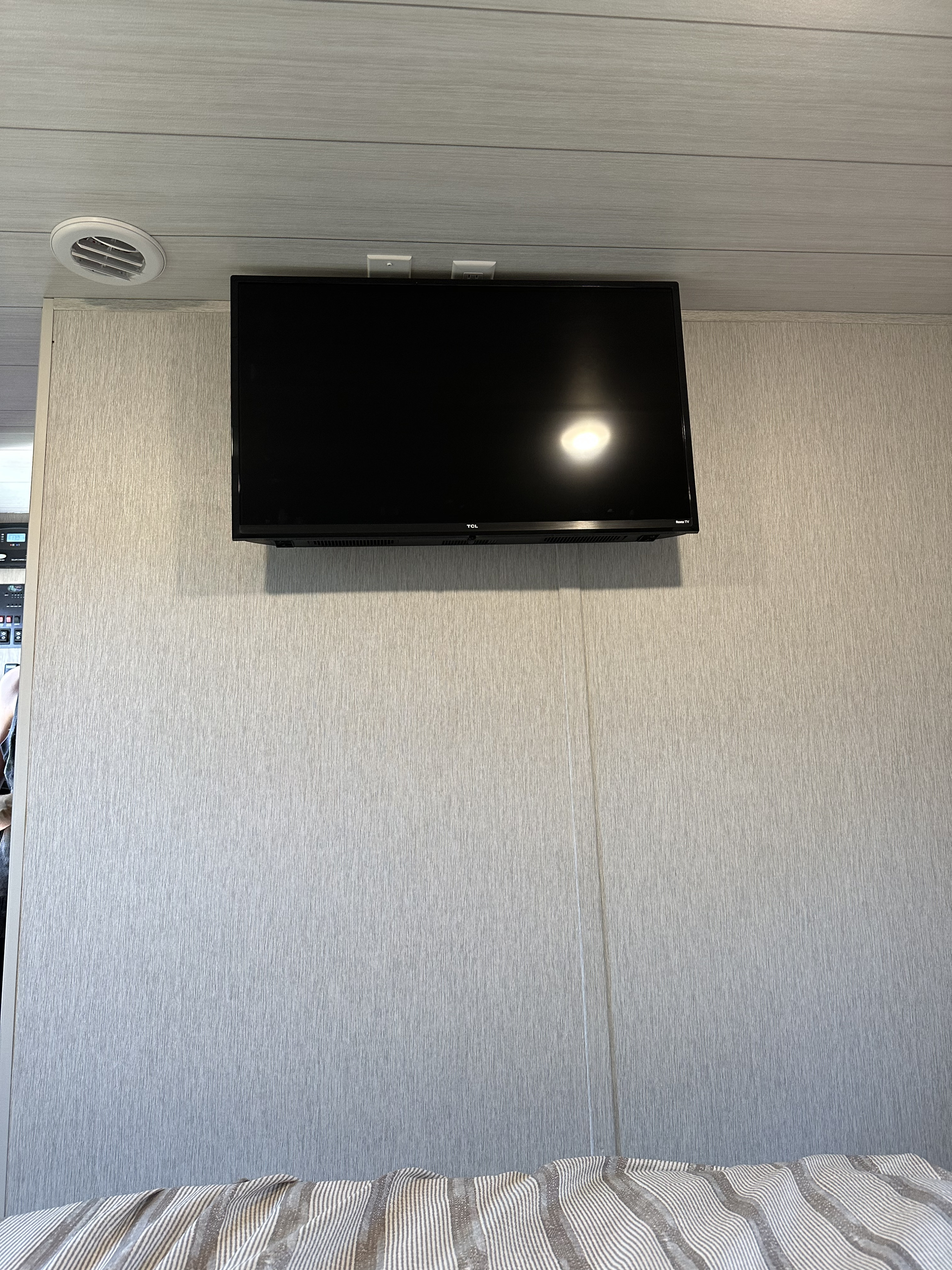 This RV rental features a flat-screen TV mounted on a textured wall, with ceiling vents and outlets, overlooking striped bedding.. Forest River Cruise Lite 2023