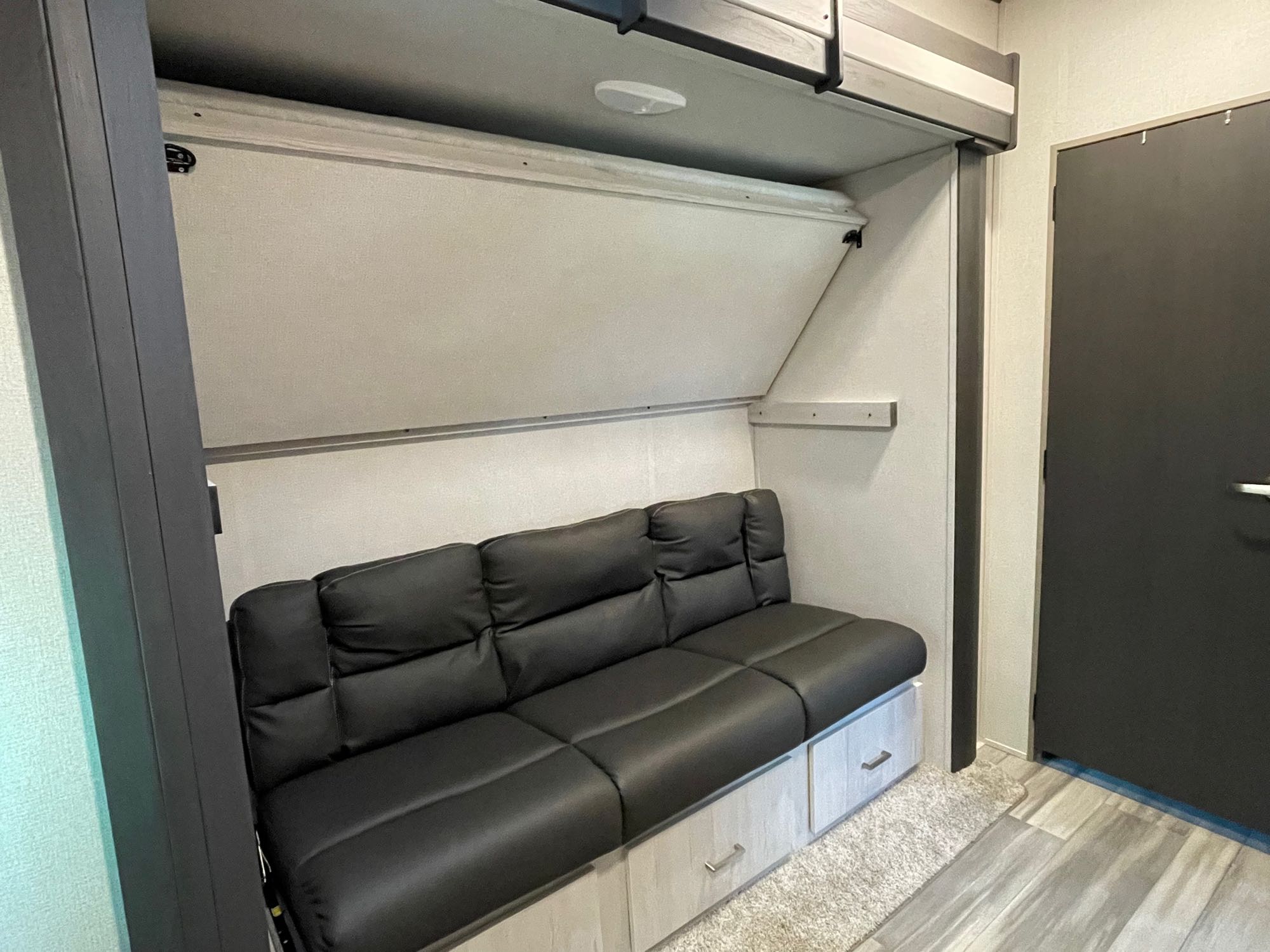 This RV rental interior shows a black sofa with storage, a fold-down bunk bed, and a dark door.. Keystone RV Sprinter 2021