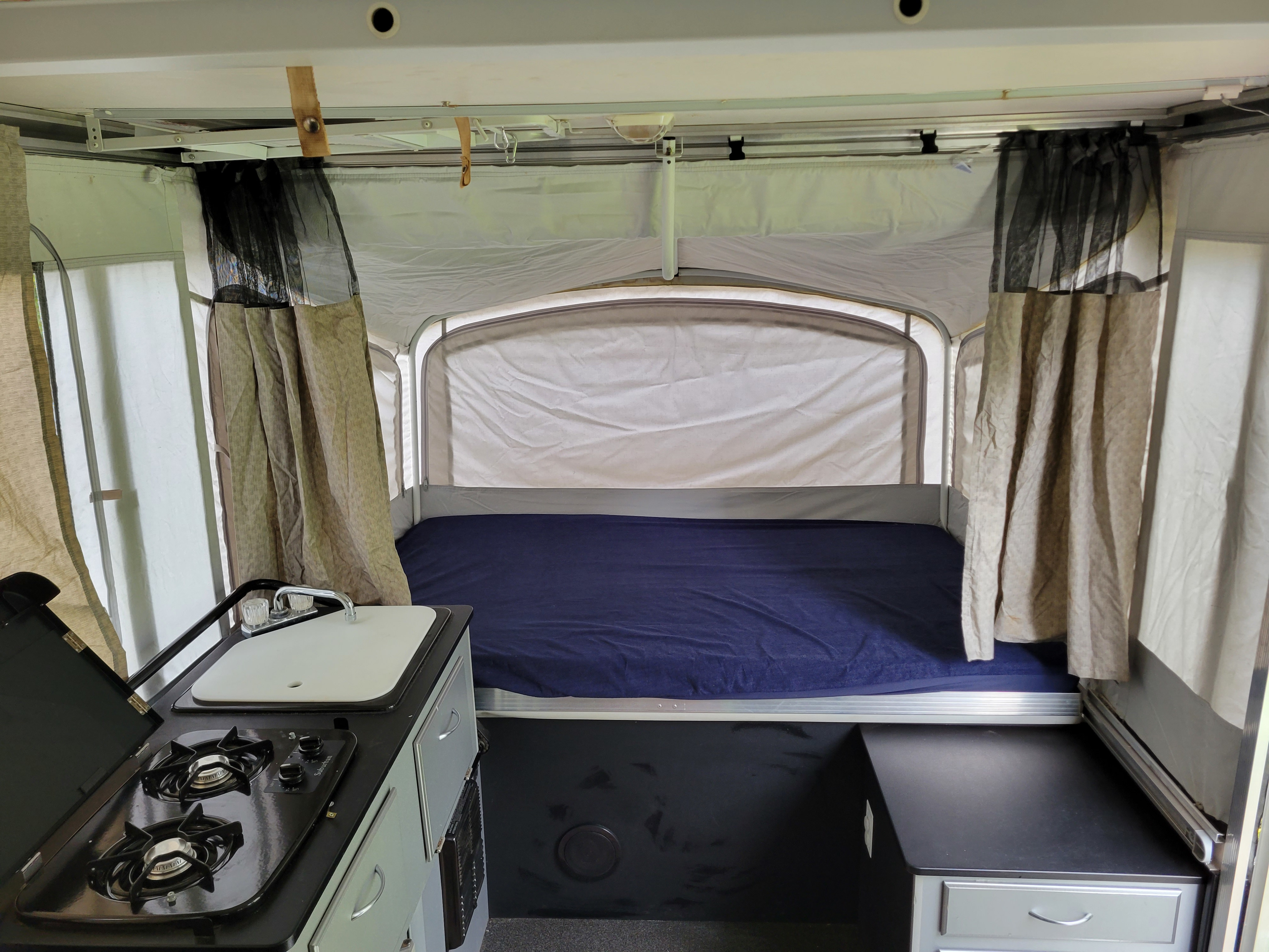 Inside this RV rental pop-up camper: a bed with blue sheets, curtains, and a compact kitchen.. Fleetwood Evolution 2008