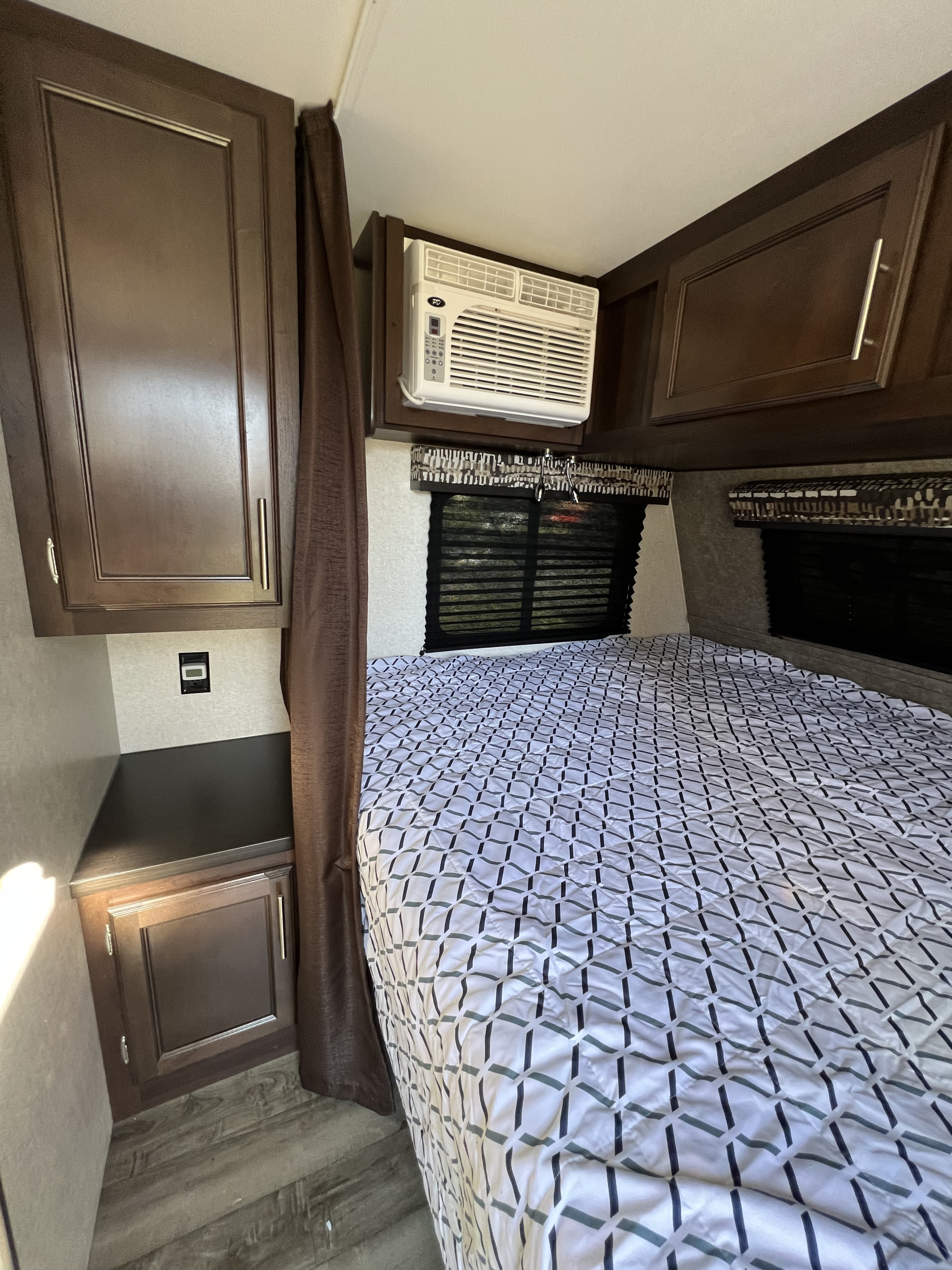 Cozy RV rental bedroom featuring dark wood cabinets, a window with blinds, an AC unit, and a patterned comforter on the bed.. Jayco Jay Flight SLX 2019