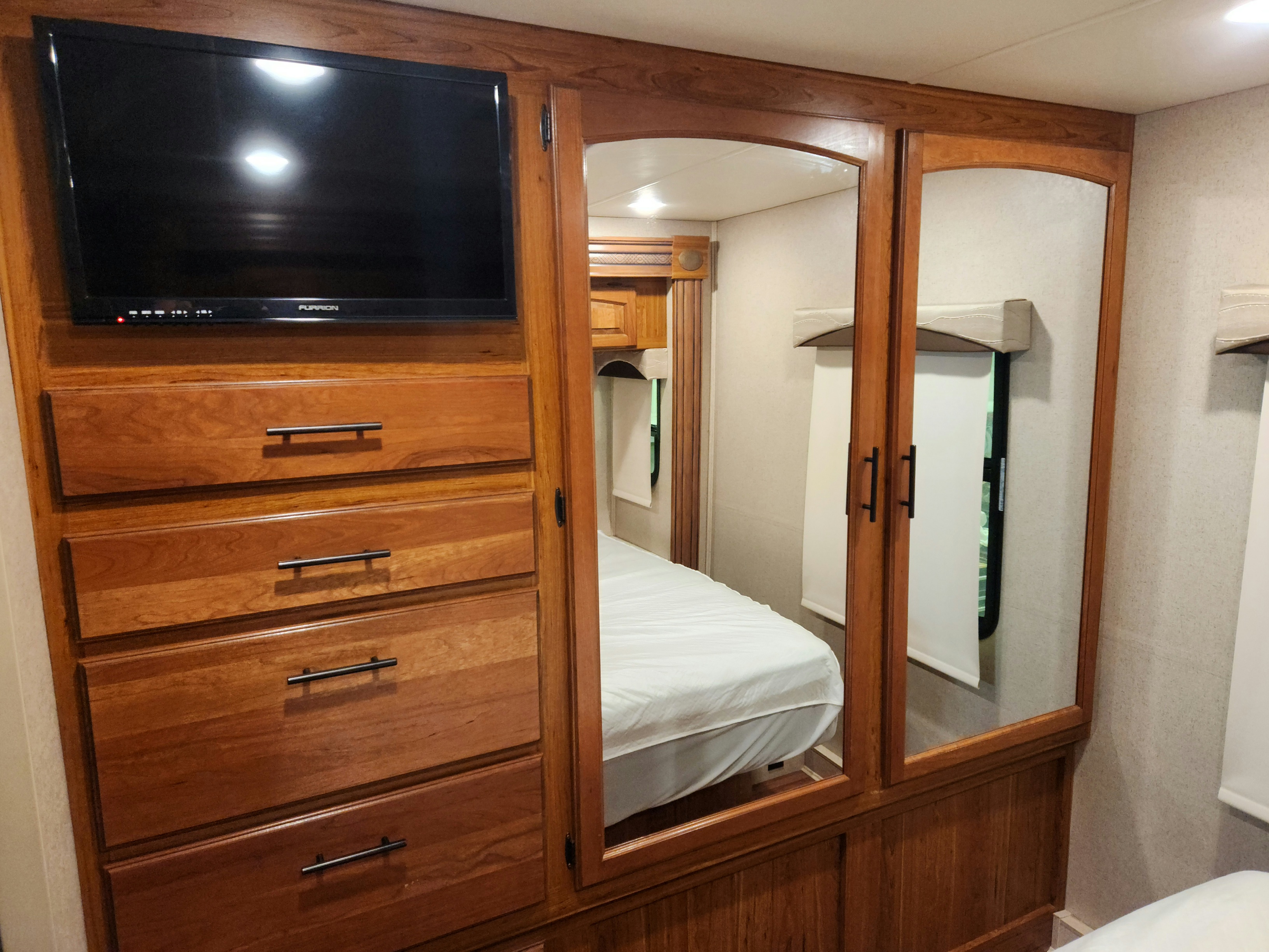 Inside an RV rental bedroom: wooden cabinetry with a TV, dresser, and mirrored wardrobe reflecting the bed.. Jayco Greyhawk 2016