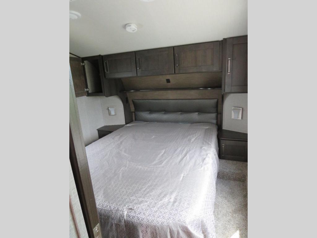 RV rental bedroom: a large bed with grey patterned cover, padded headboard, dark wood overhead cabinets & nightstands.. Grand Design Reflection 2022