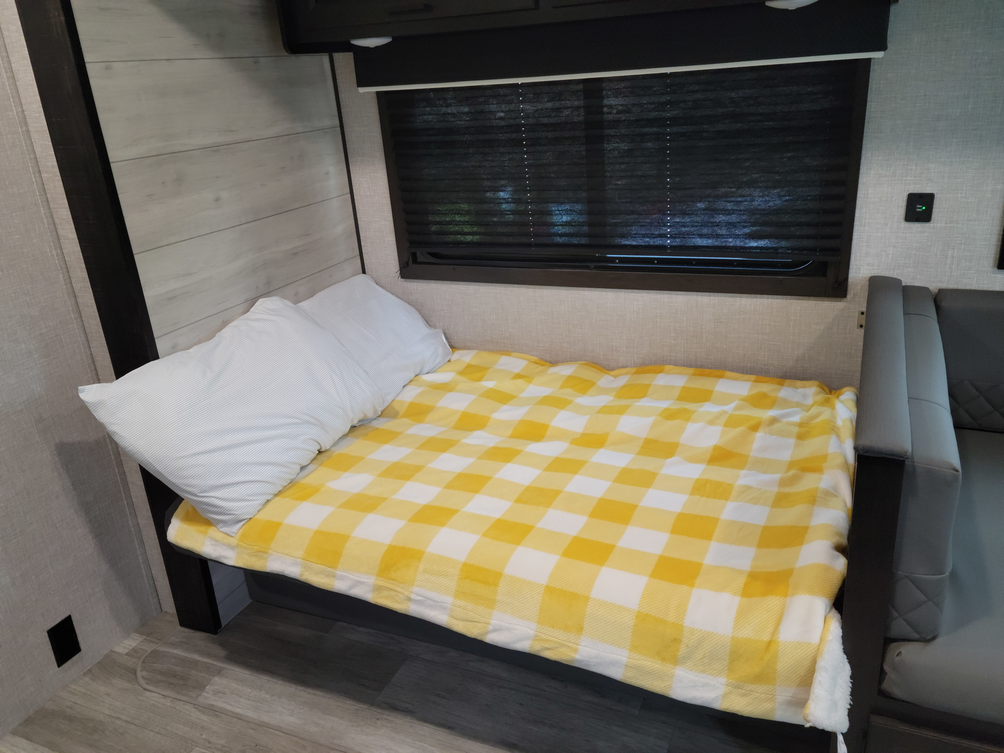 Cozy RV rental sleeping area featuring a yellow plaid blanket, white pillow, window, and nearby couch.. Jayco Jay Flight 2023