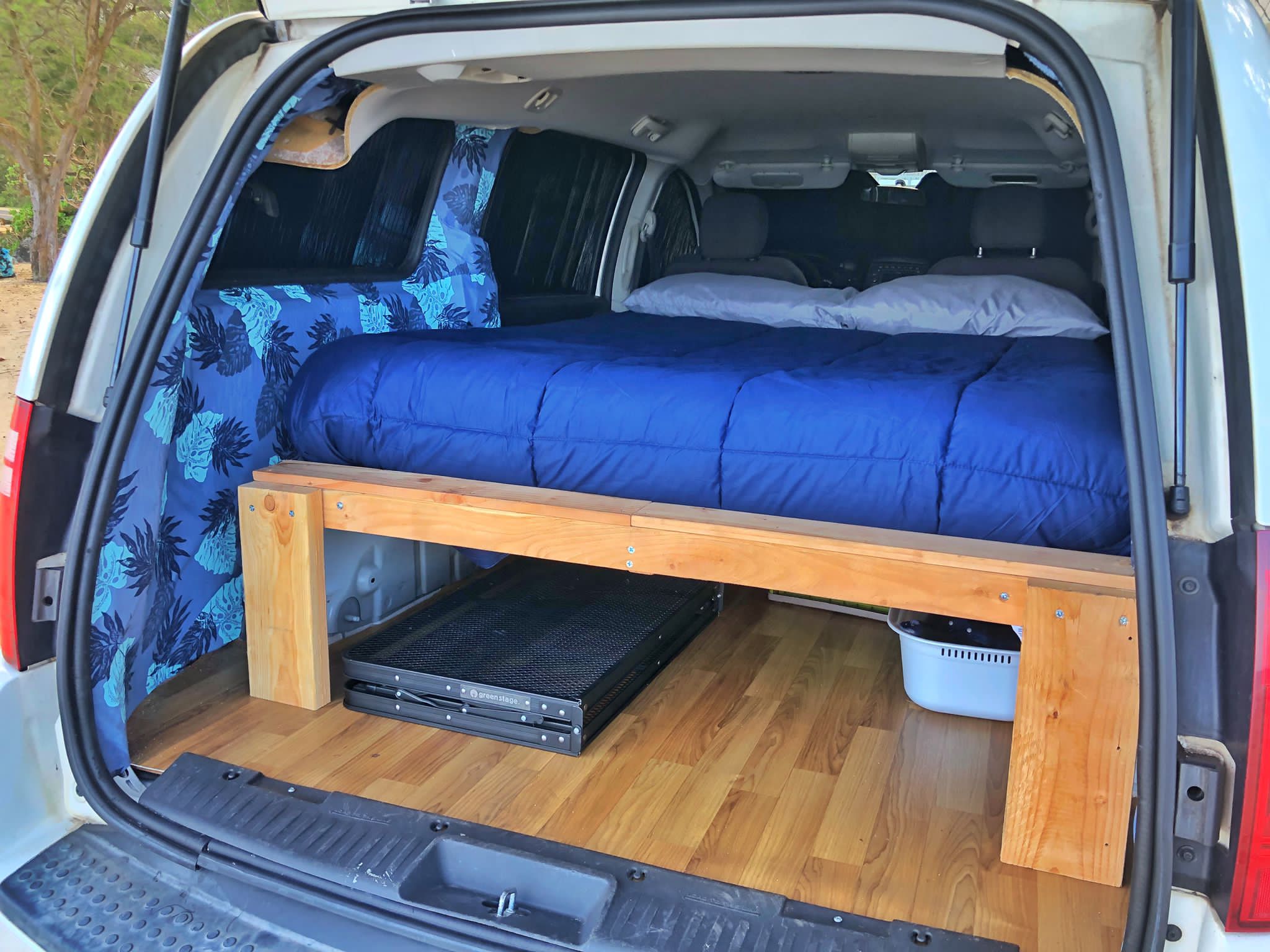 This RV rental shows a cozy van conversion: a bed with blue bedding, under-bed storage, & tropical curtains.. Dodge Grand Caravan 2010