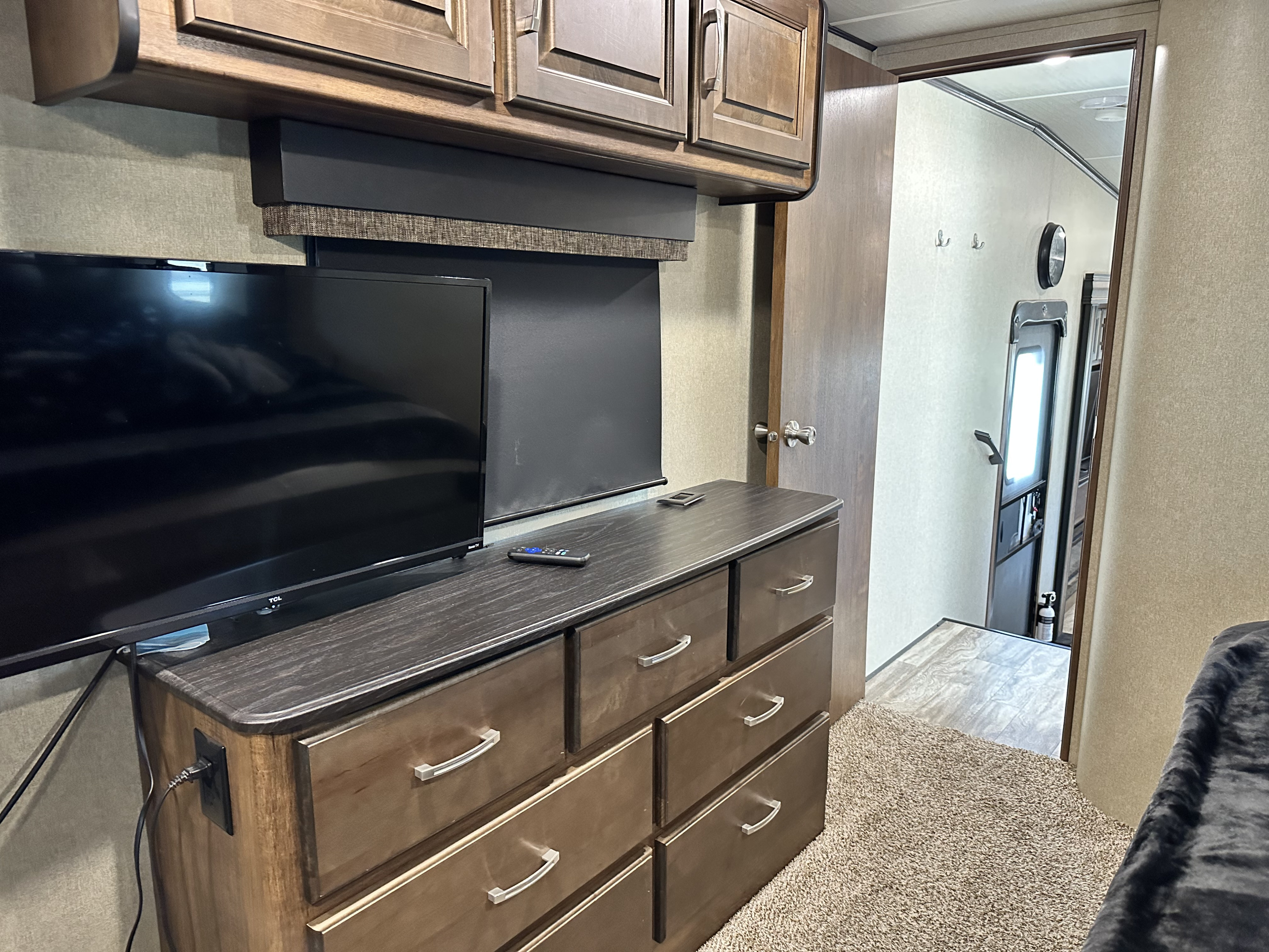 Inside an RV rental bedroom: a TV sits on a dark wood dresser with drawers, topped by wooden cabinets. A door opens to a hallway.. Grand Design Reflection 2020