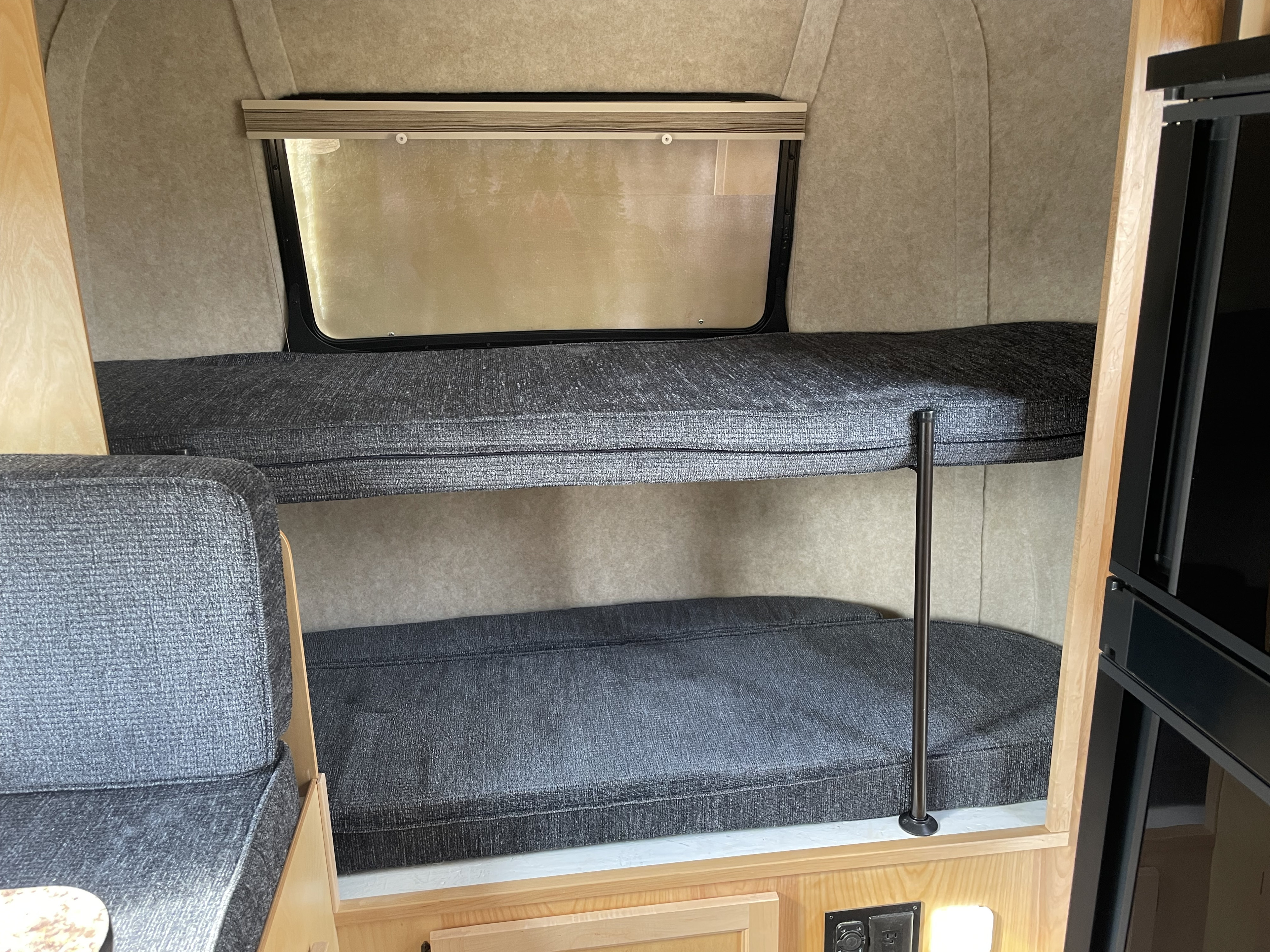 RV rental interior featuring stacked bunk beds with grey cushions. A window is above the top bunk, with wooden trim and power outlets below.. Scamp Deluxe 2023