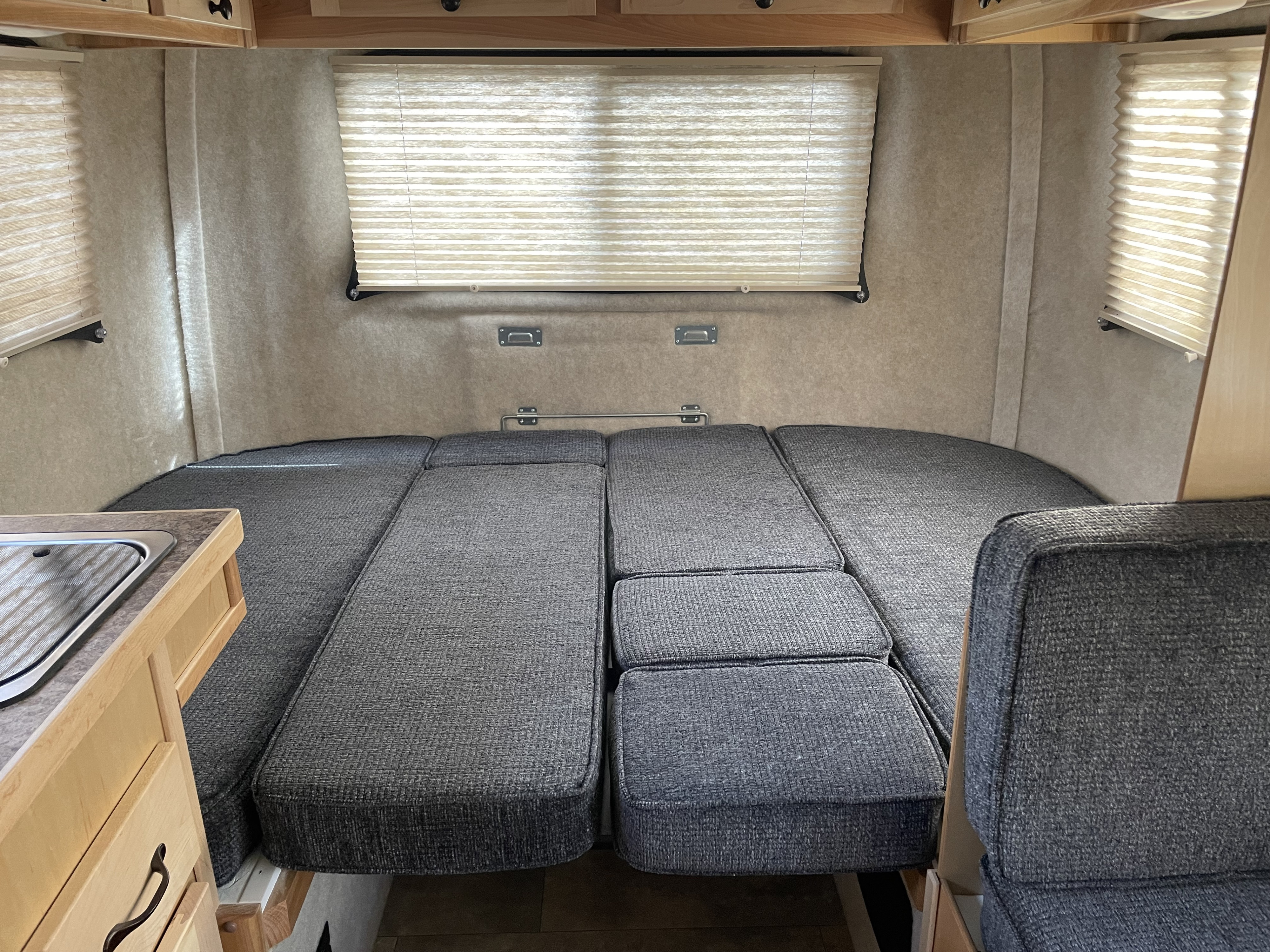 Inside this **rv rental**: a cozy space with a large gray upholstered bed/seating area, pleated window blinds, and a compact sink.. Scamp Deluxe 2023