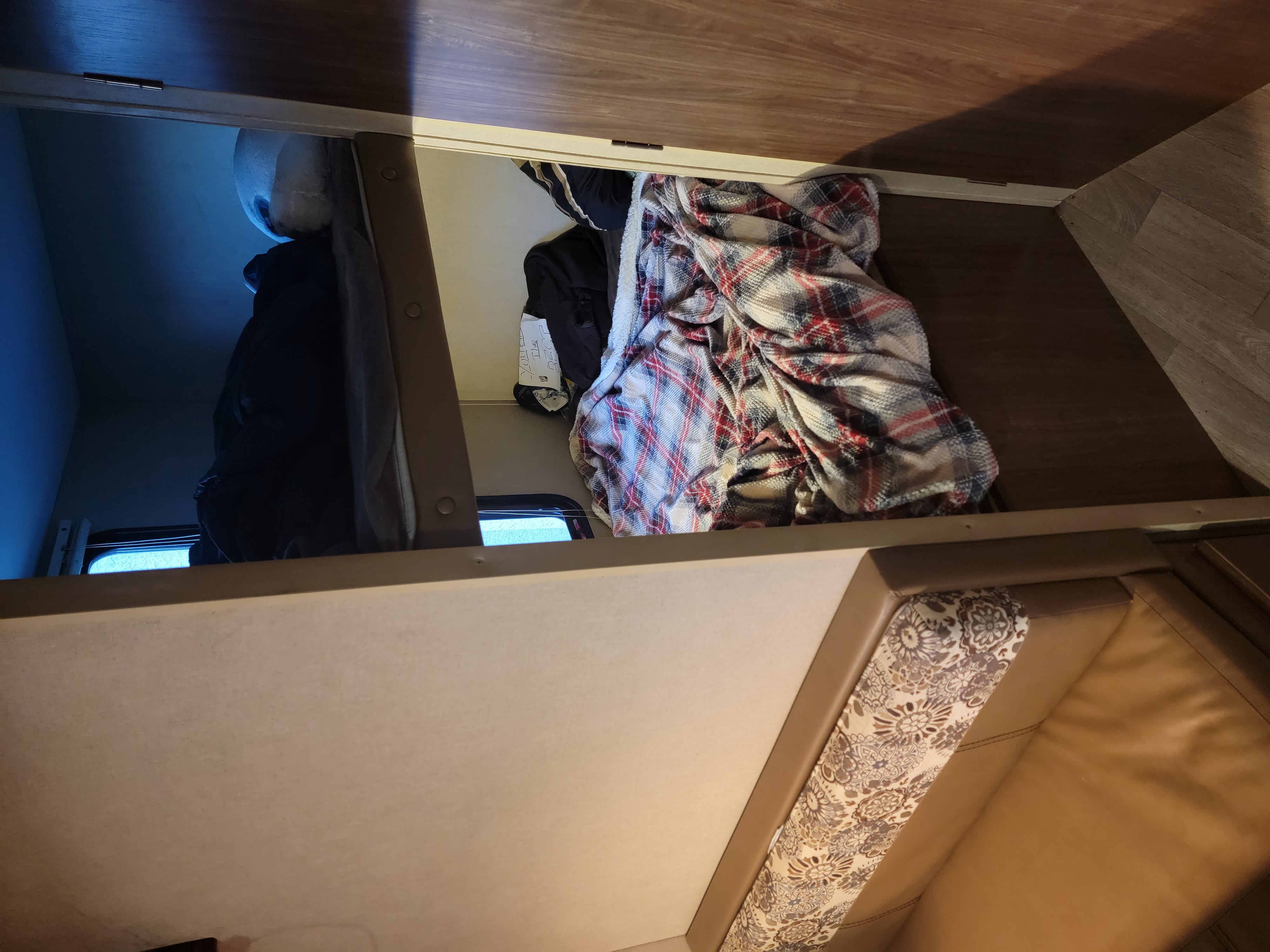 View into an RV rental overhead compartment with a plaid blanket, clothing, and window, highlighting camper storage.. Dutchmen Coleman Lantern 2016