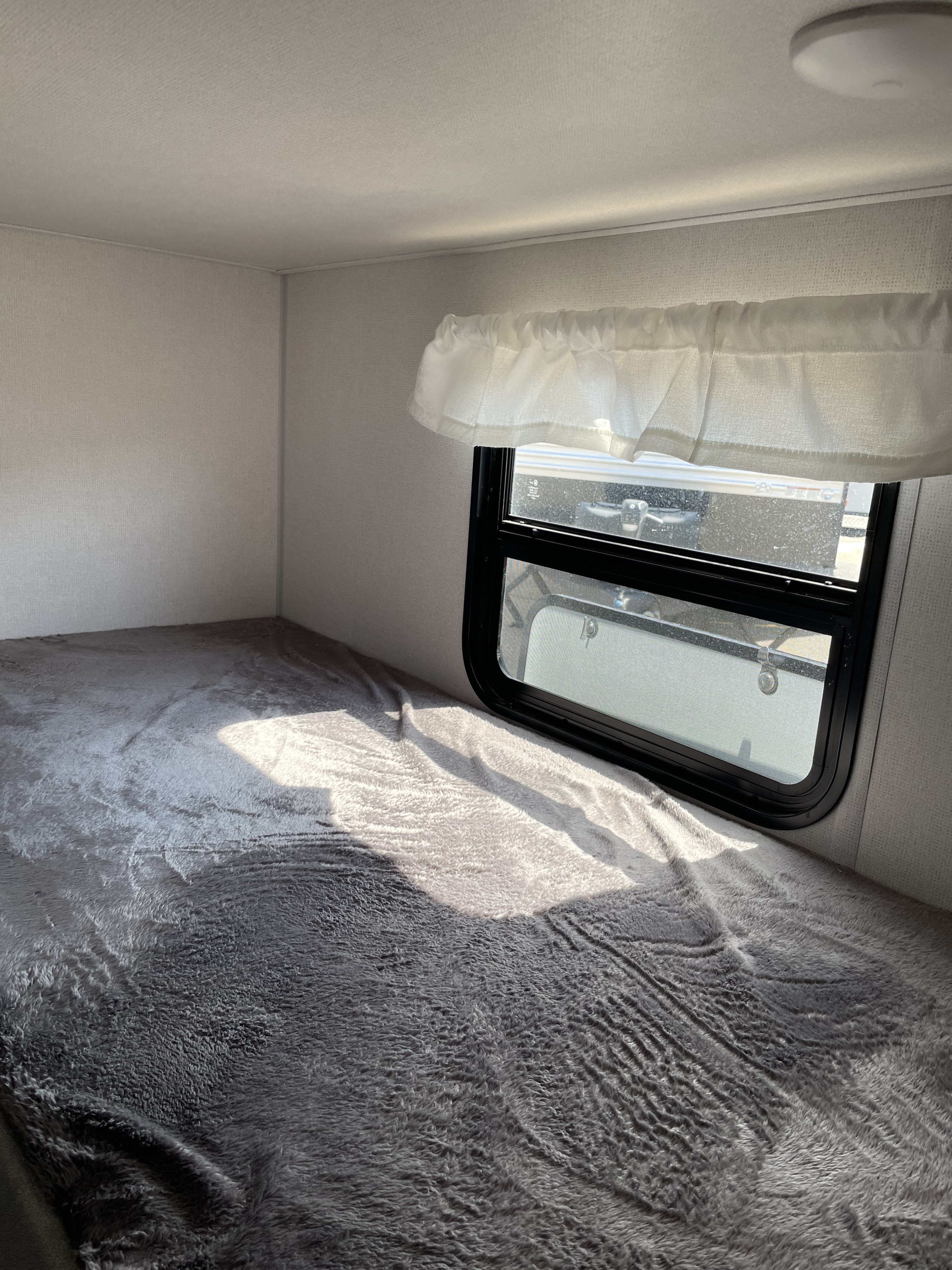 Cozy RV rental sleeping area features a plush gray bed under a window with a sheer white valance, illuminated by sunlight.. Avenger LT 2022