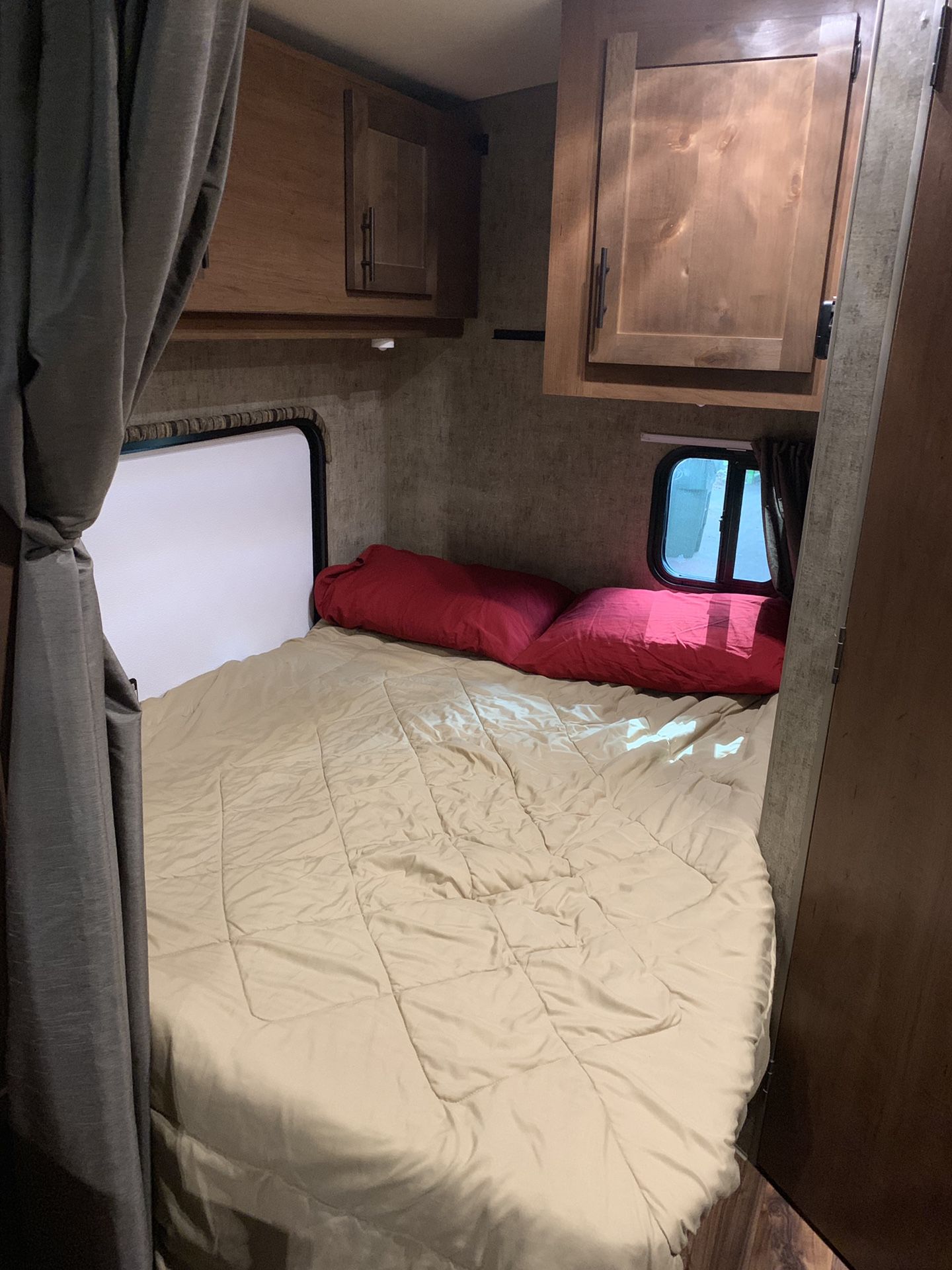 This rv rental interior shows a compact bedroom with a beige bed, red pillows, two windows, and wooden overhead cabinets.. Outdoors RV Blackrock 2017