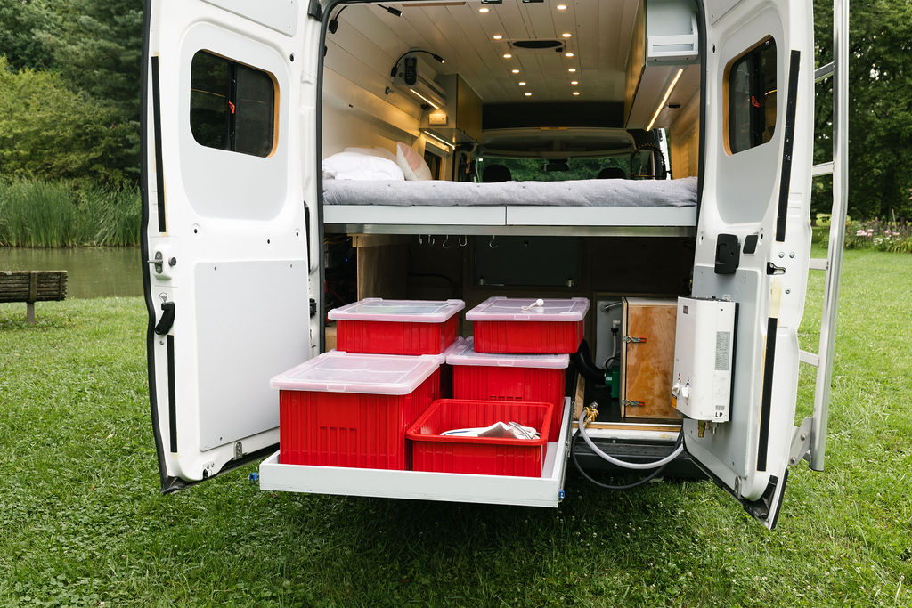 RV rental camper van interior: rear doors reveal a bed, pull-out storage with red bins, and external water heater.. Dodge Promaster 159 2019