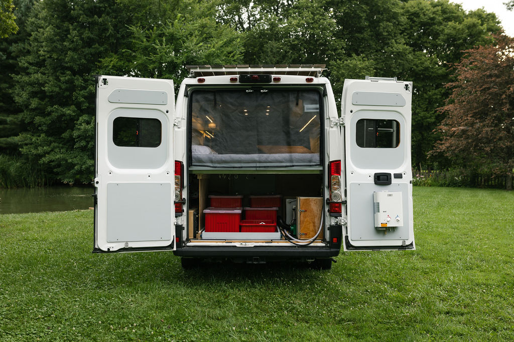 Rear of an RV rental camper van with open doors, showing a bed, storage, and external water heater, ready for travel.. Dodge Promaster 159 2019