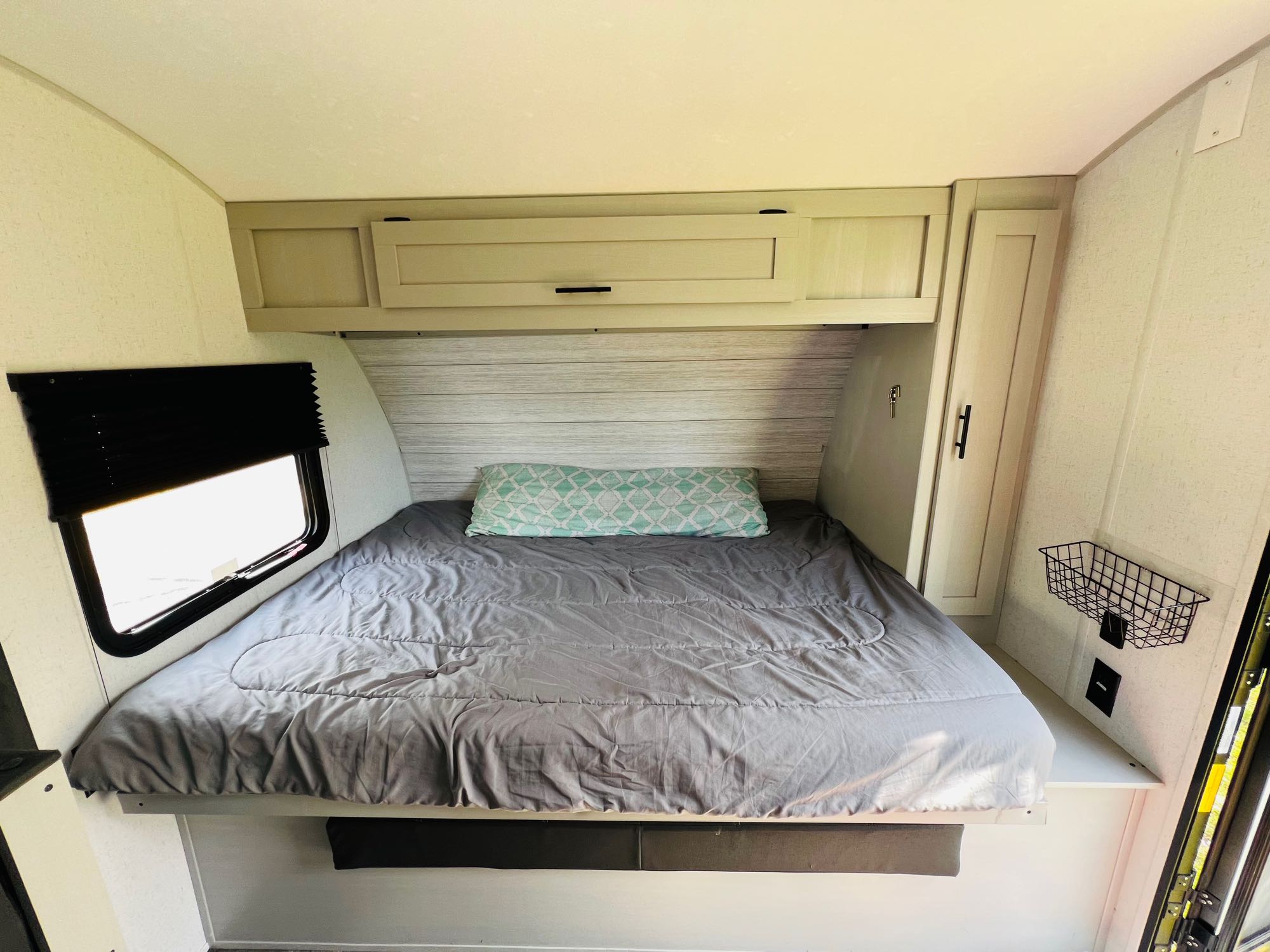 This clean RV rental bedroom features a gray bed with a patterned pillow, overhead cabinets, and a window with blinds.. Forest River Wildwood 2022