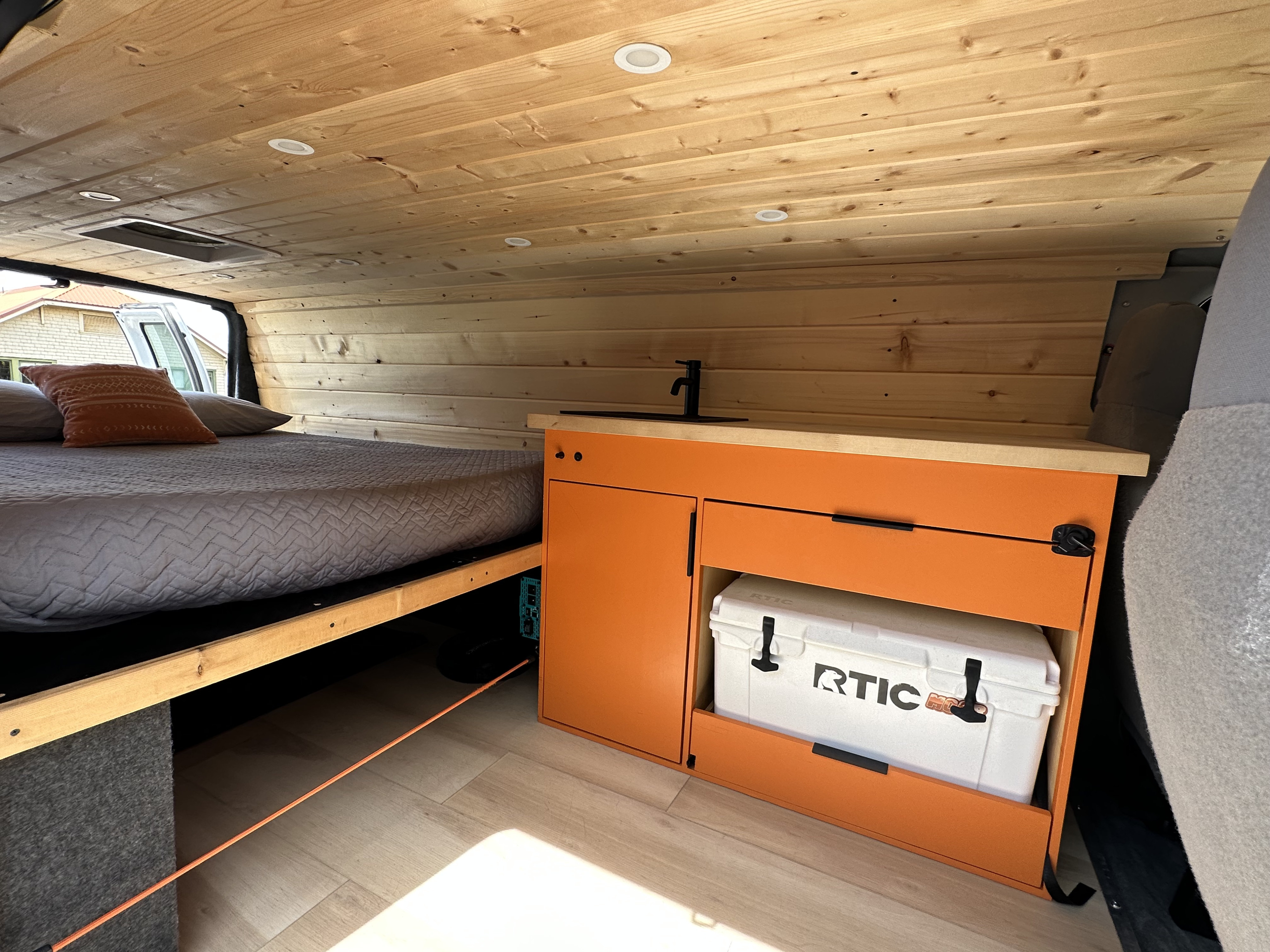 This RV rental showcases a custom interior with a bed, wood paneling, and an orange kitchen cabinet storing an RTIC cooler.. Ford Extended E350 2011