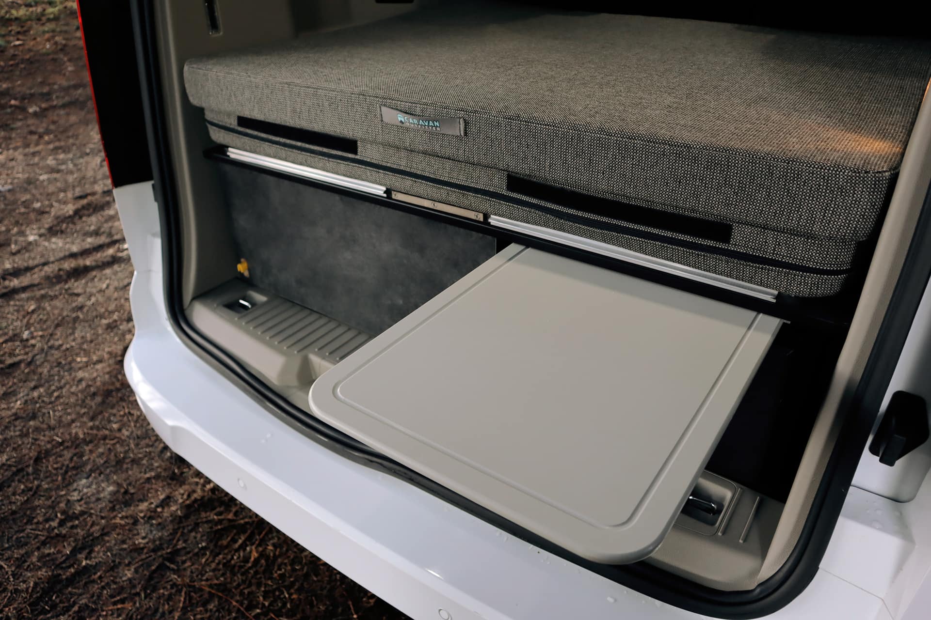 Inside an RV rental: a compact, modular bed with "Caravan Master" branding, and a grey pull-out table extended from beneath.. Ford Transit Connect Wagon 2022
