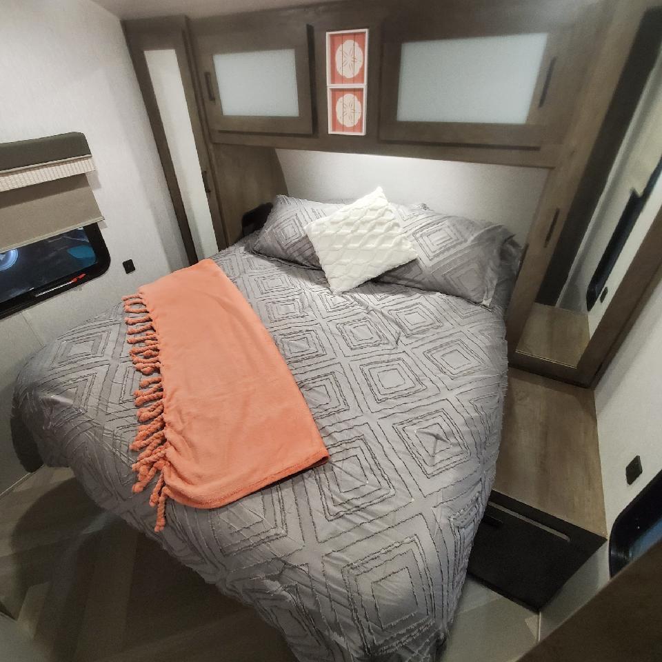 Compact and cozy, this RV rental bedroom has a gray bed with a peach throw, overhead cabinets, and a window.. Forest River Salem 2022