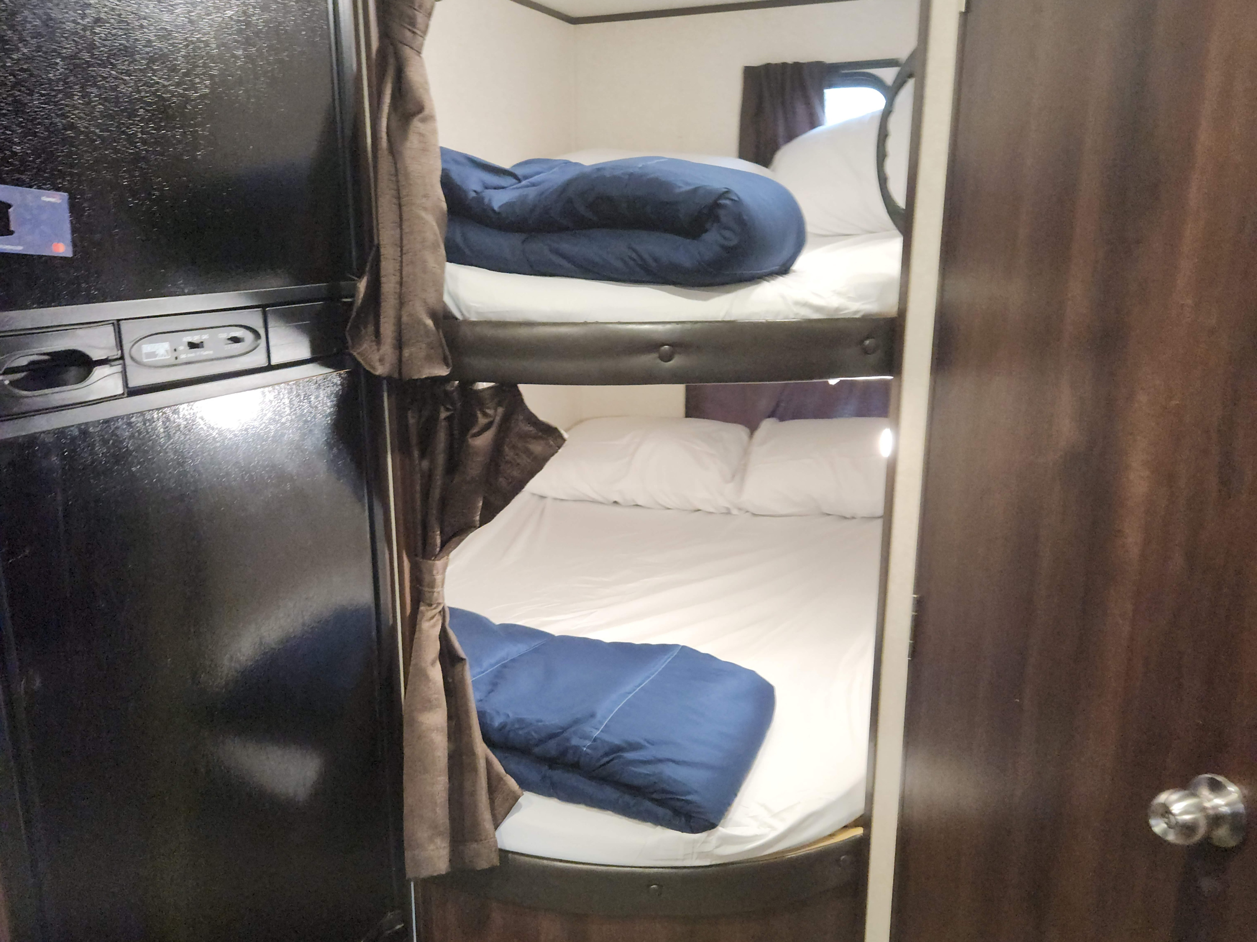Cozy RV rental interior with a compact refrigerator on the left and stacked bunk beds, featuring white sheets and blue comforters, on the right.. Jayco Jay Flight SLX 2019