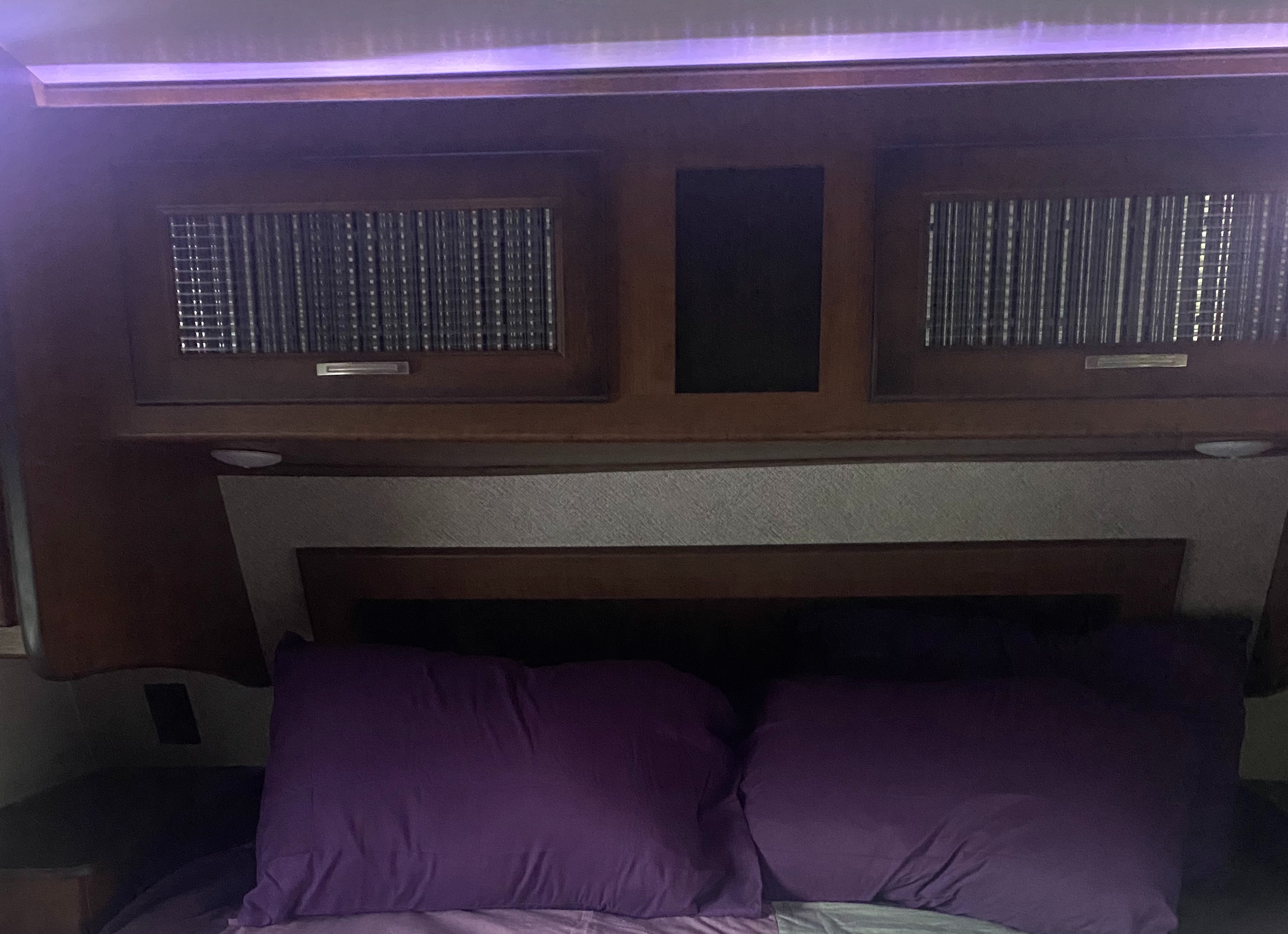 Comfortable RV rental bedroom featuring dark wood overhead cabinetry, a padded headboard, and inviting purple pillows.. Heartland RVs Elkridge 2018
