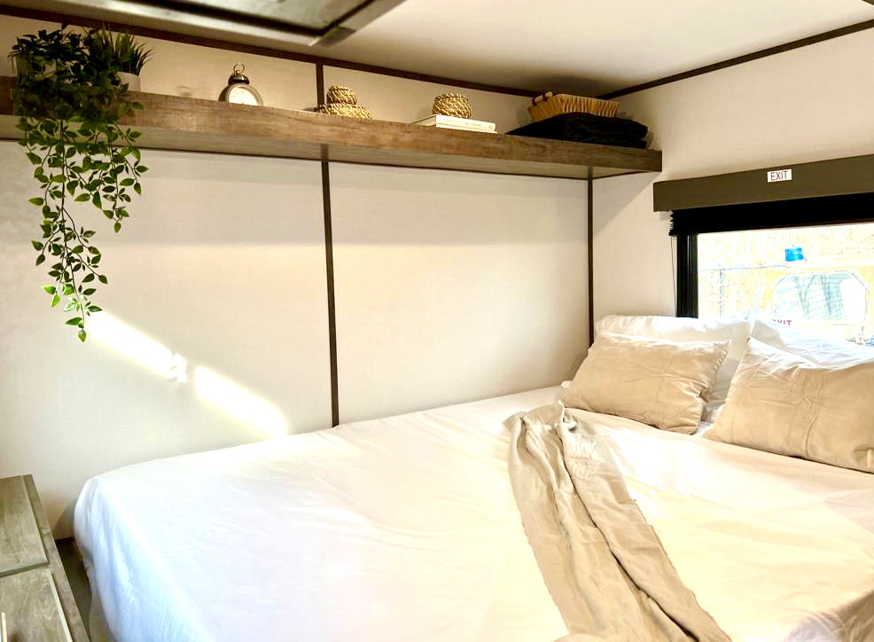 RV rental bedroom: comfortable bed with white linens, decorative wooden shelf, and a bright window with an "EXIT" sign.. Riverside RV Retro 165 2022