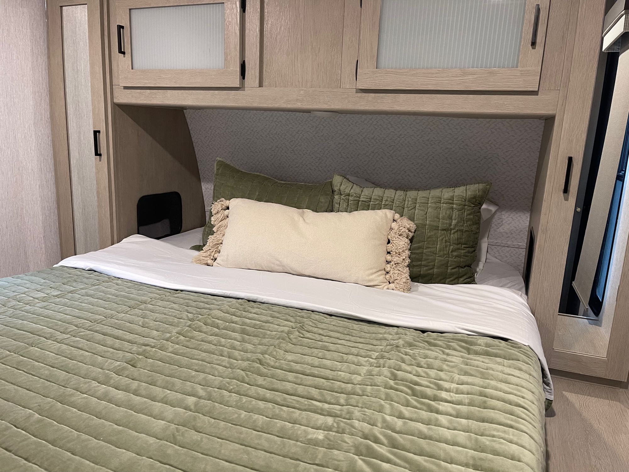 Inside an RV rental: a neatly made bed with green comforter, cozy pillows, and light wood cabinetry with storage.. Forest River Wildwood 32BHDS Platinum 2023