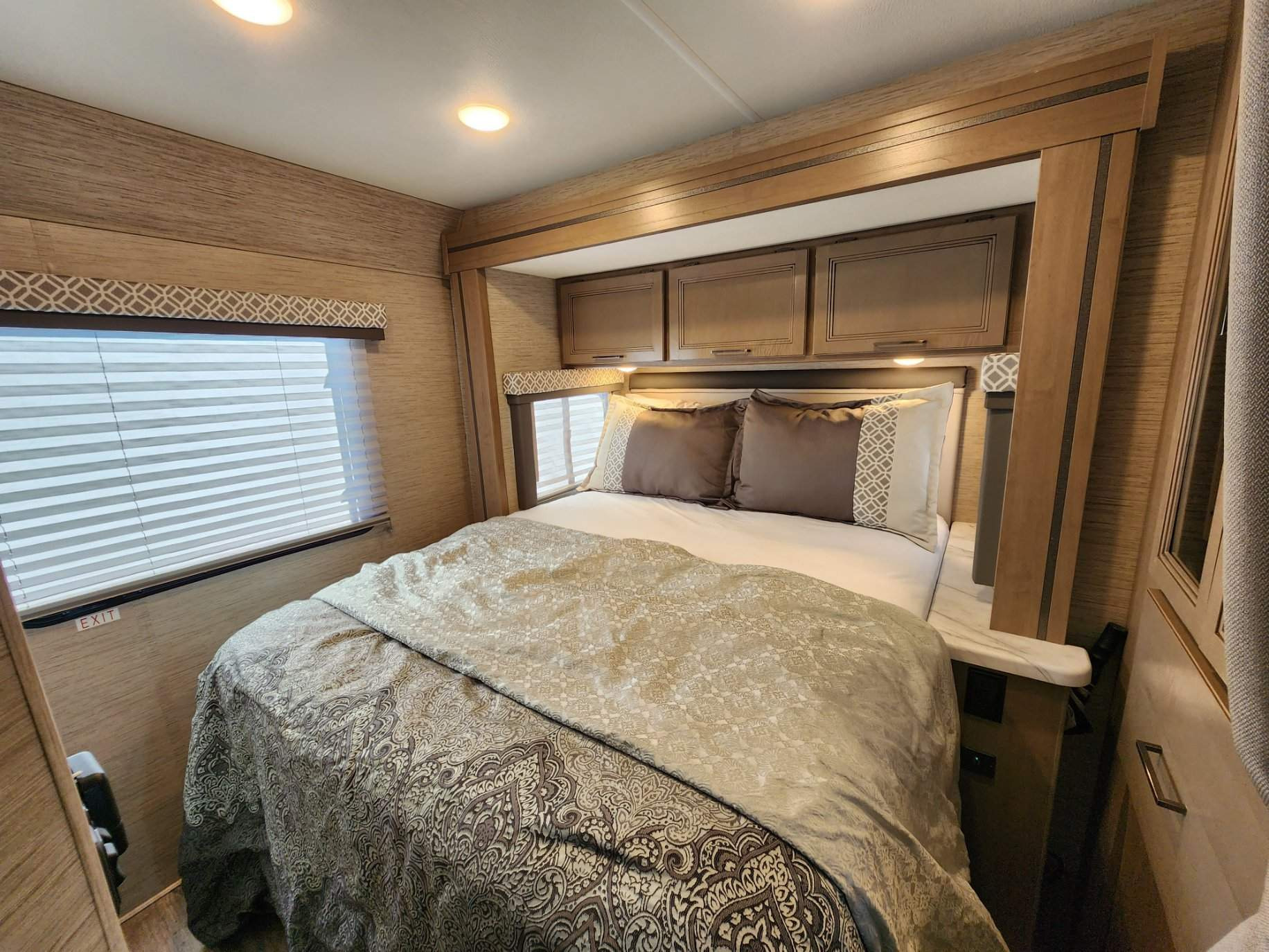 Inside a cozy RV rental bedroom: a large bed with decorative bedding, overhead cabinets, and a window with blinds.. Thor COLEMAN 2023
