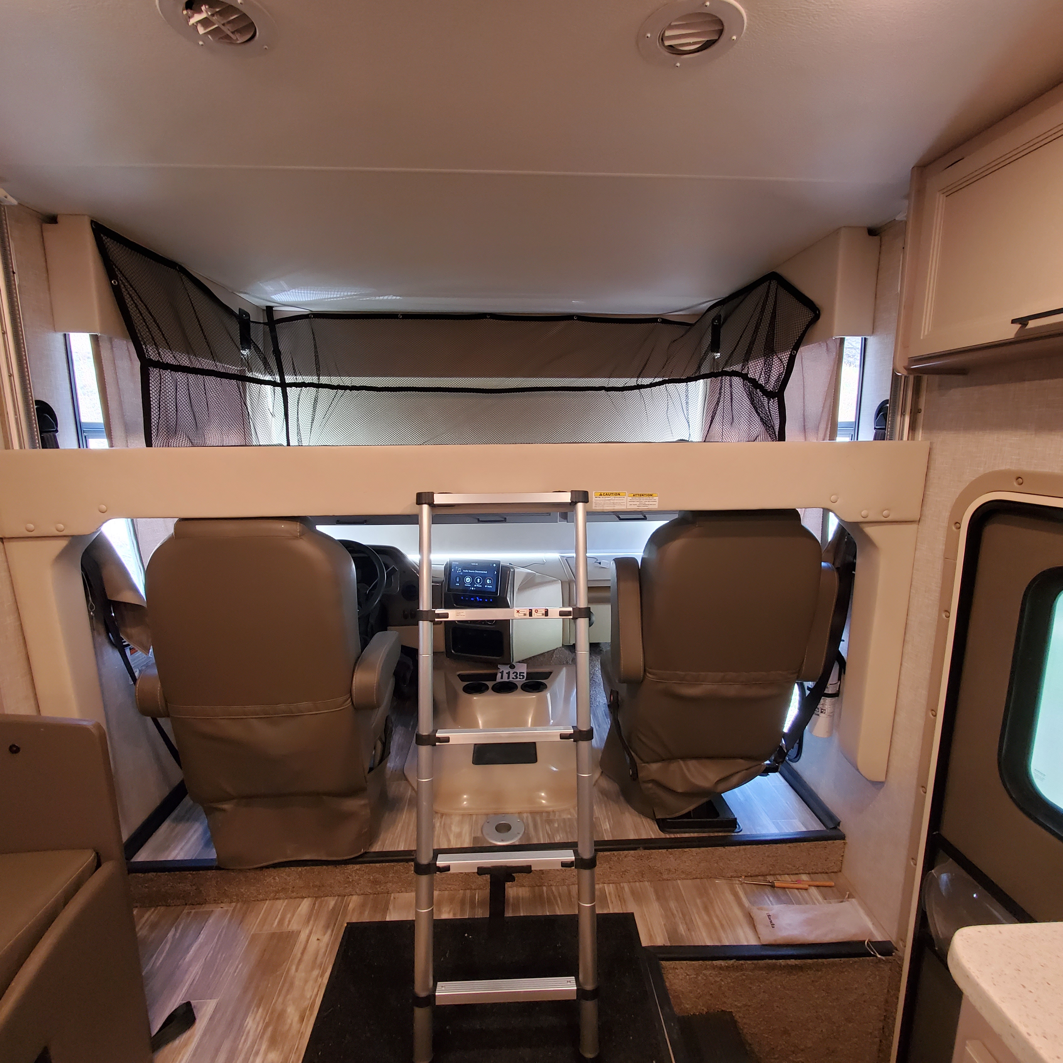 Inside an rv rental: a front view of the cockpit with driver seats & overhead bunk bed, featuring a ladder and safety netting.. Thor Hurricane 2022