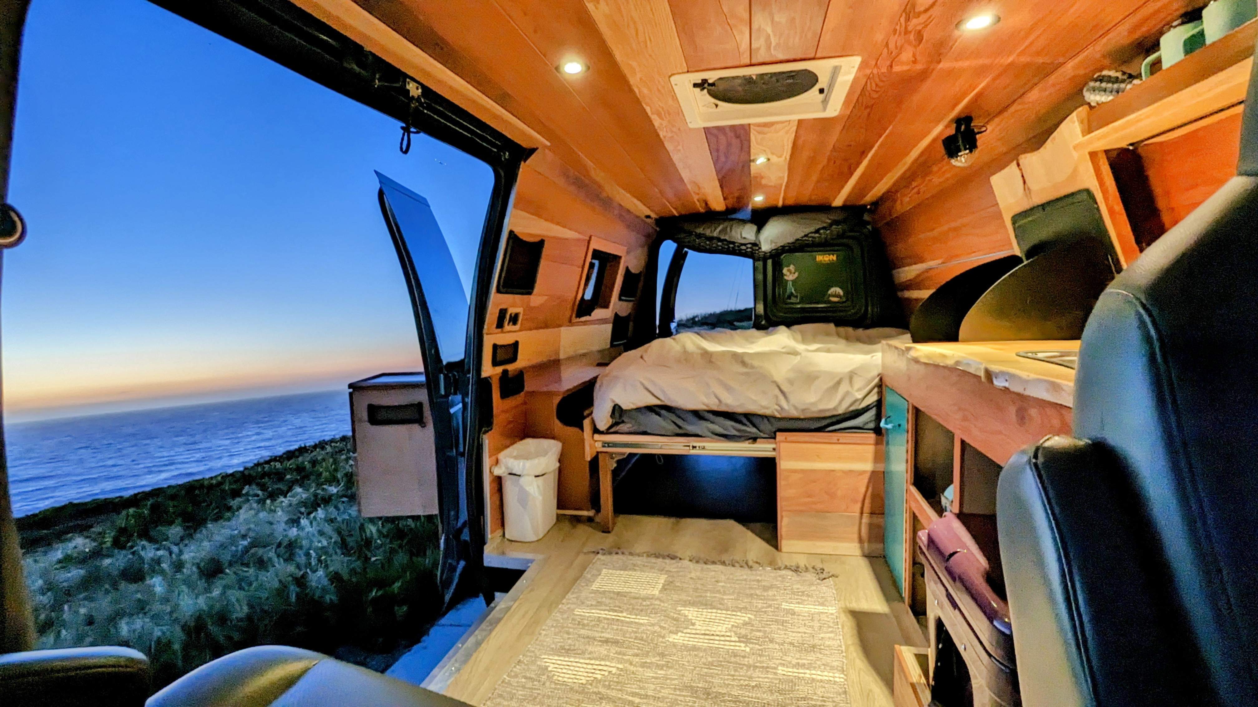 Inside this custom wood RV rental, a cozy bed & kitchenette face an open door revealing a serene ocean view at sunset.. Ford Econoline 2009