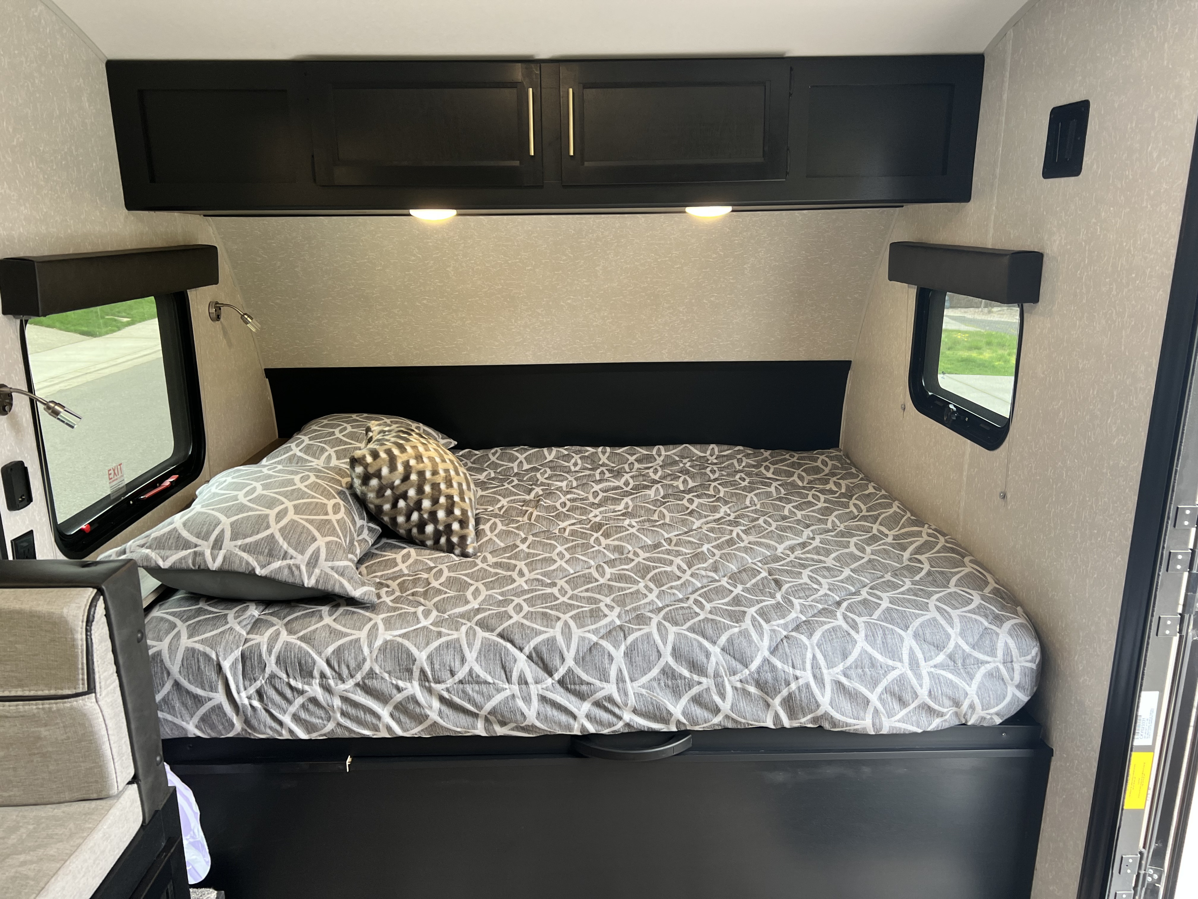 An RV rental's cozy bedroom features a gray patterned bed, black overhead cabinets, reading lights, and side windows.. Cherokee Pup Black label #3 17jgbl 2023