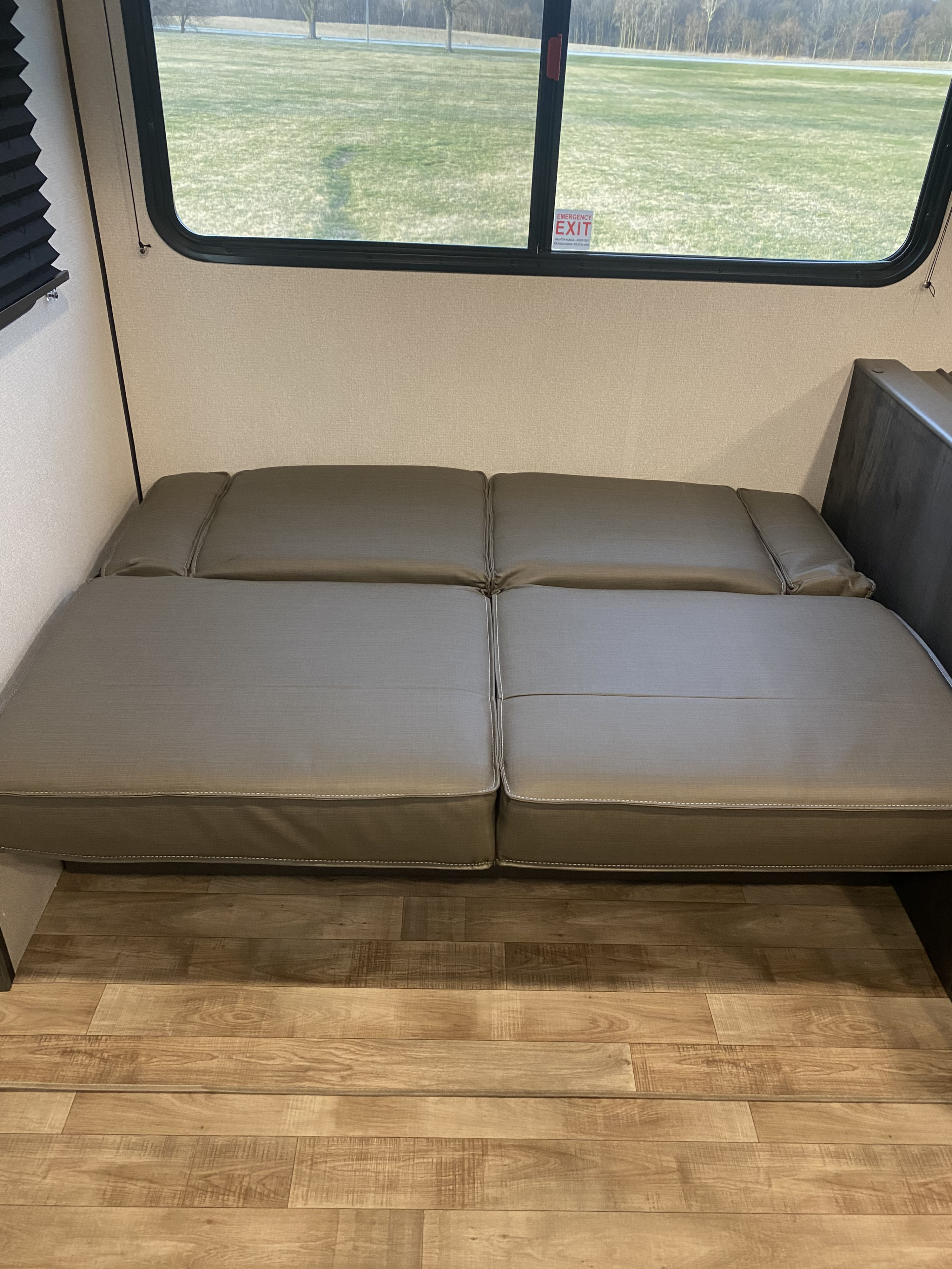 Inside an RV rental: a plush grey sofa converts to a bed, beside an emergency exit window and laminate flooring.. Keystone hideout 29dfs 2023