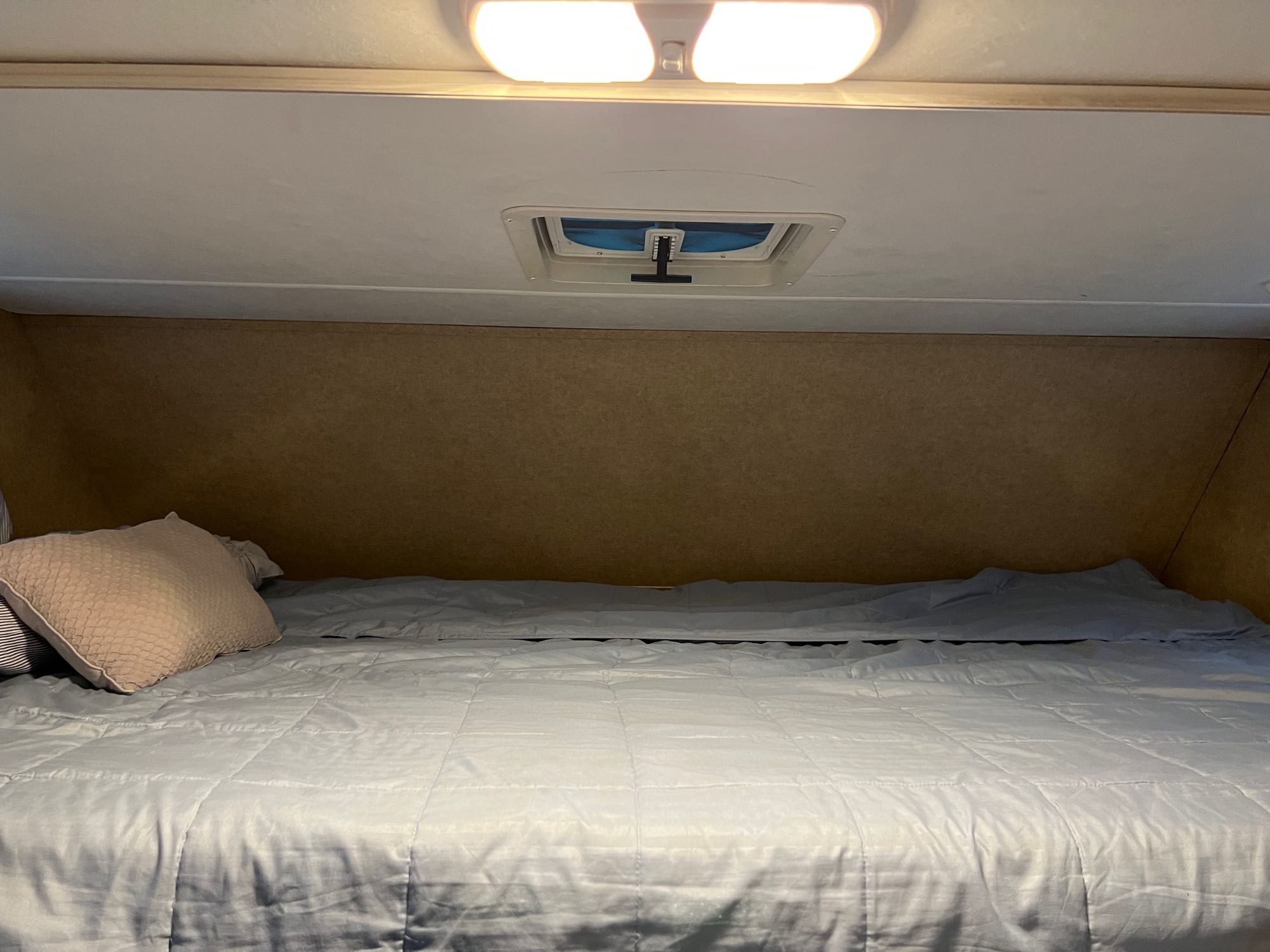 Cozy RV rental sleeping area: a bed with light blue bedding and a beige pillow, illuminated by overhead lights and a vent.. Thor Majestic 2016