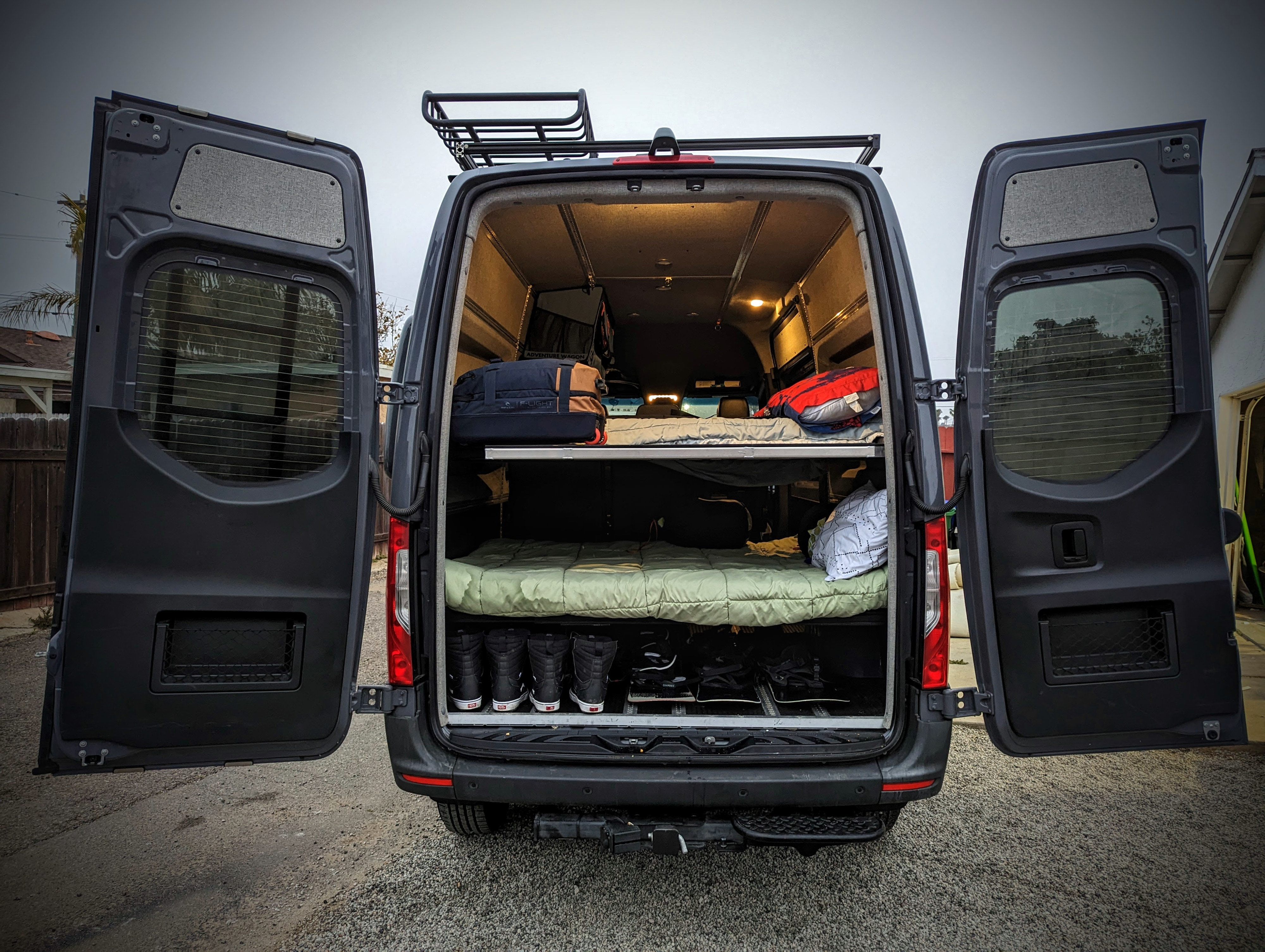 Rear view of a spacious RV rental van with open doors revealing bunk beds, organized gear storage, and a roof rack for travel.. Mercedes-Benz sprinter 2021