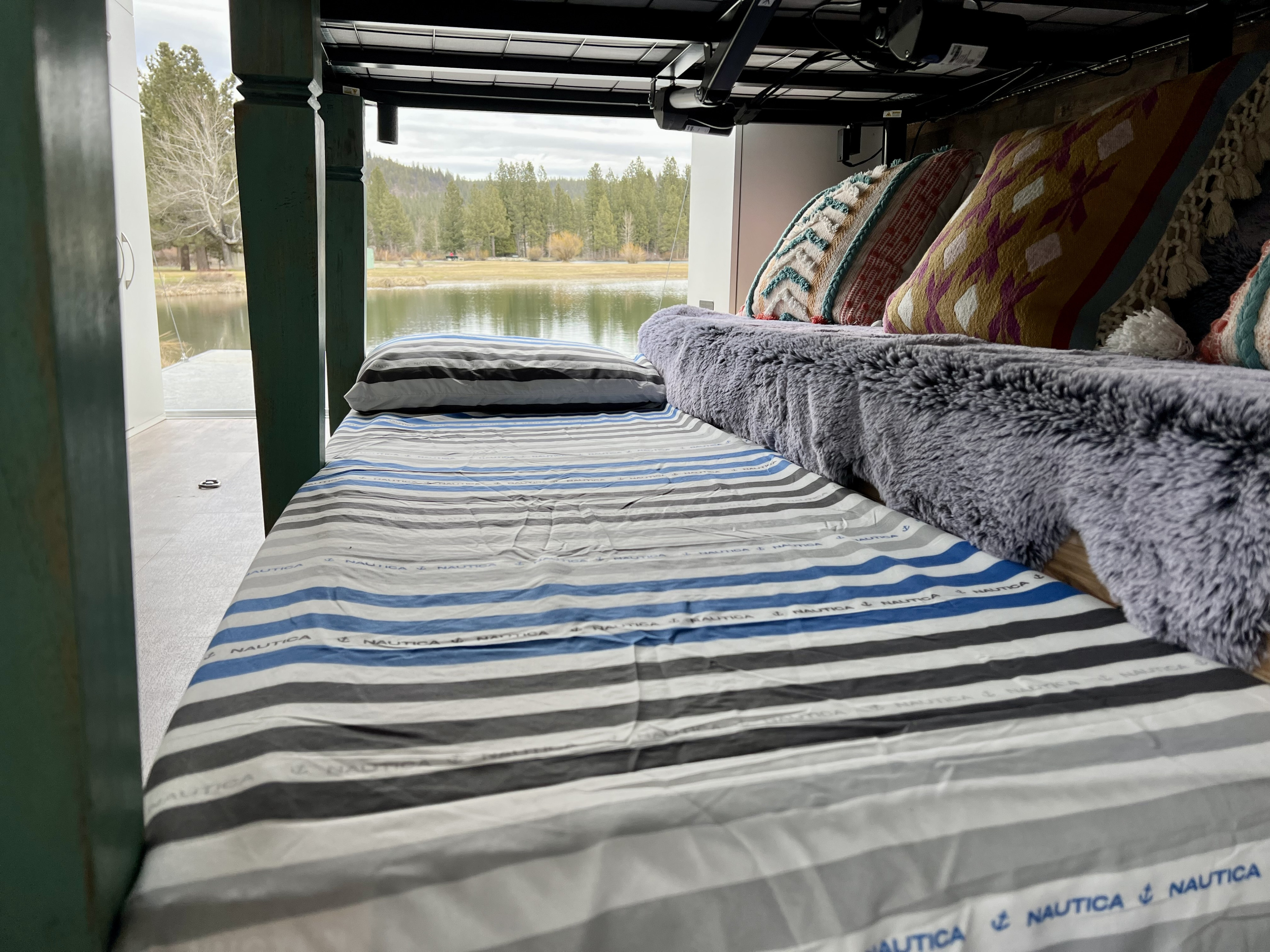 Experience tranquil travel from this RV rental bed with striped sheets, decorative pillows, and a scenic lake view.. Cargo Pro Stealth 2021