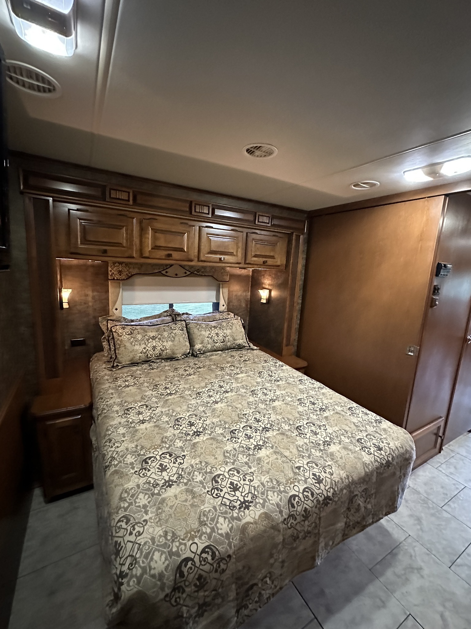 Comfortable RV rental bedroom featuring a patterned bed, overhead cabinets, window, and tiled flooring.. Allegro Allegro Motorhome 2012