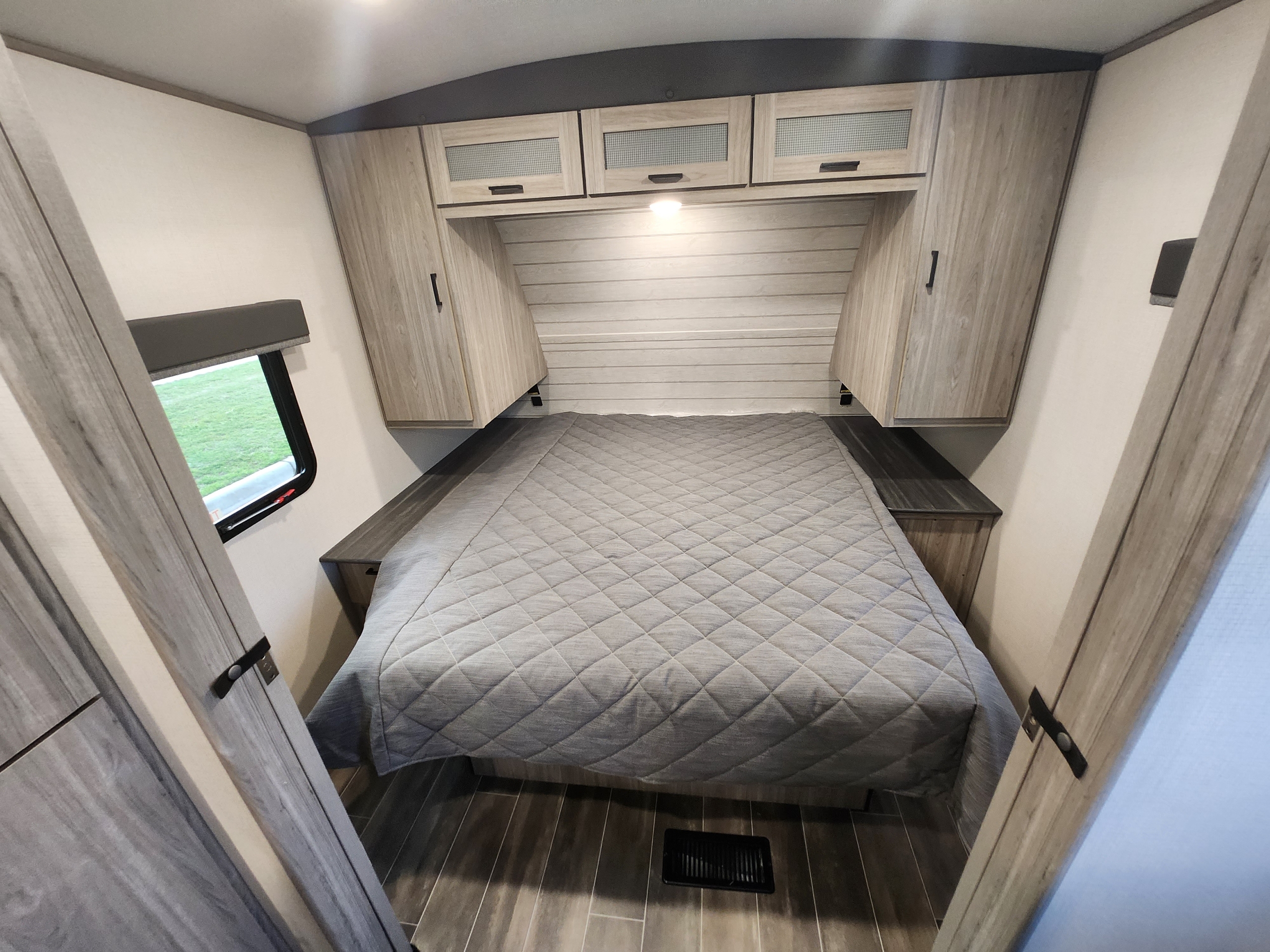 Interior view of an RV rental bedroom showing a bed with a grey comforter, built-in storage, and a window.. Keystone RV Passport GT Ultra Lite 2023