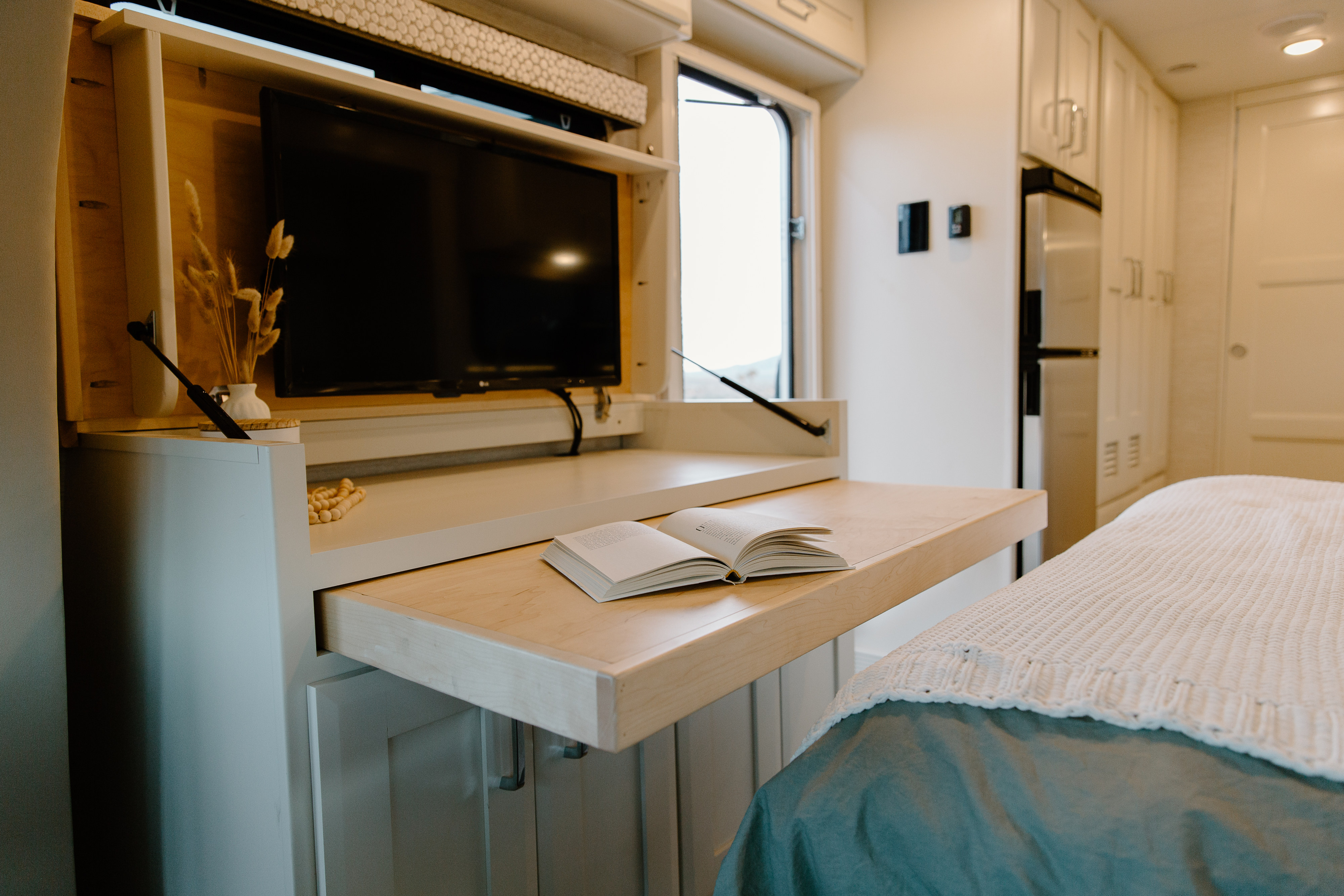 Bright RV rental interior shows a clever pull-out desk with TV, cozy bed, and white cabinetry. Compact living.. Tiffin Motorhomes Wayfarer 25 JW 2021