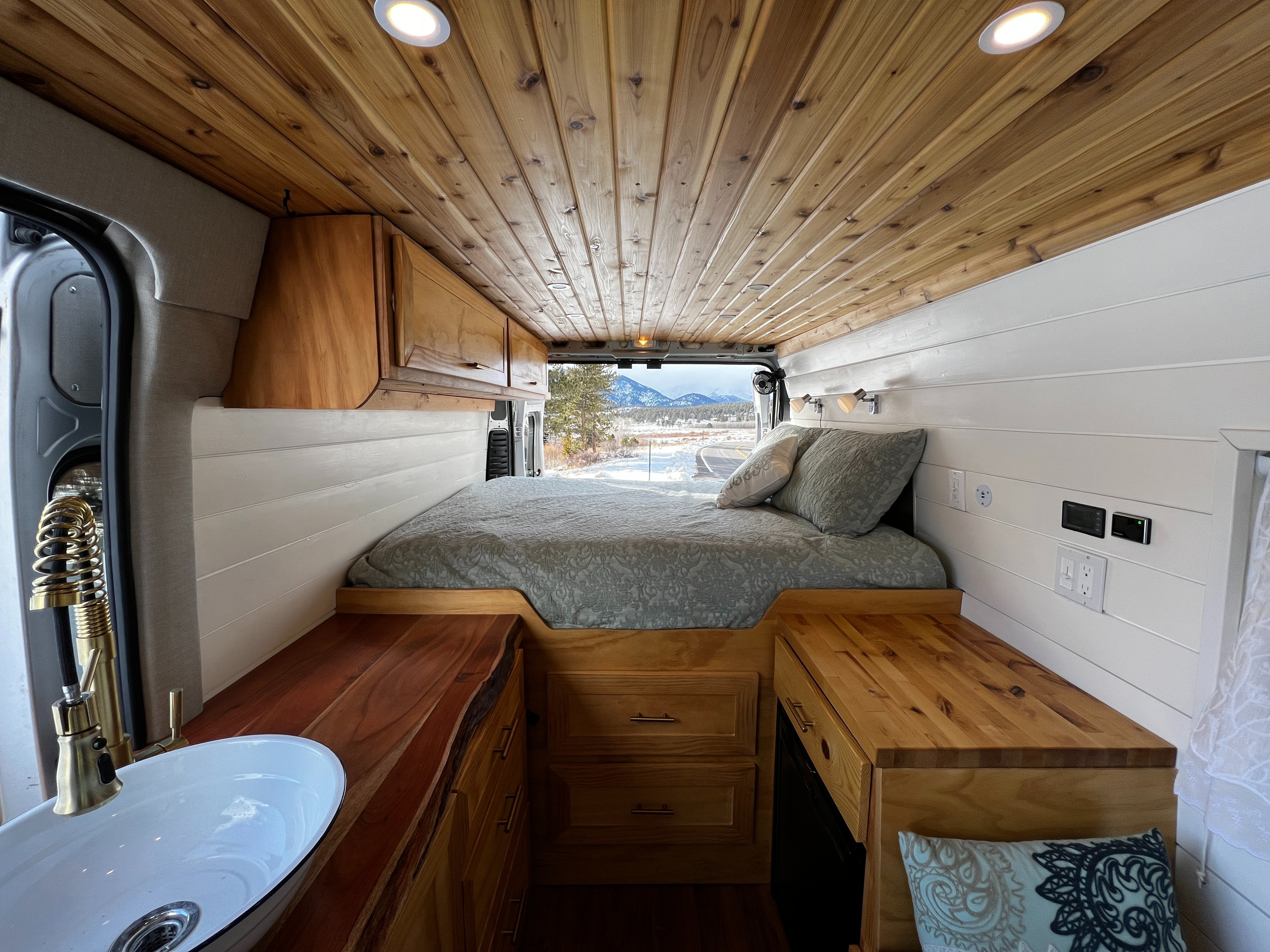 Cozy RV rental interior: wood-paneled ceiling, white walls, comfortable bed, compact kitchen sink. Mountain views outside.. Ram Promaster 2019