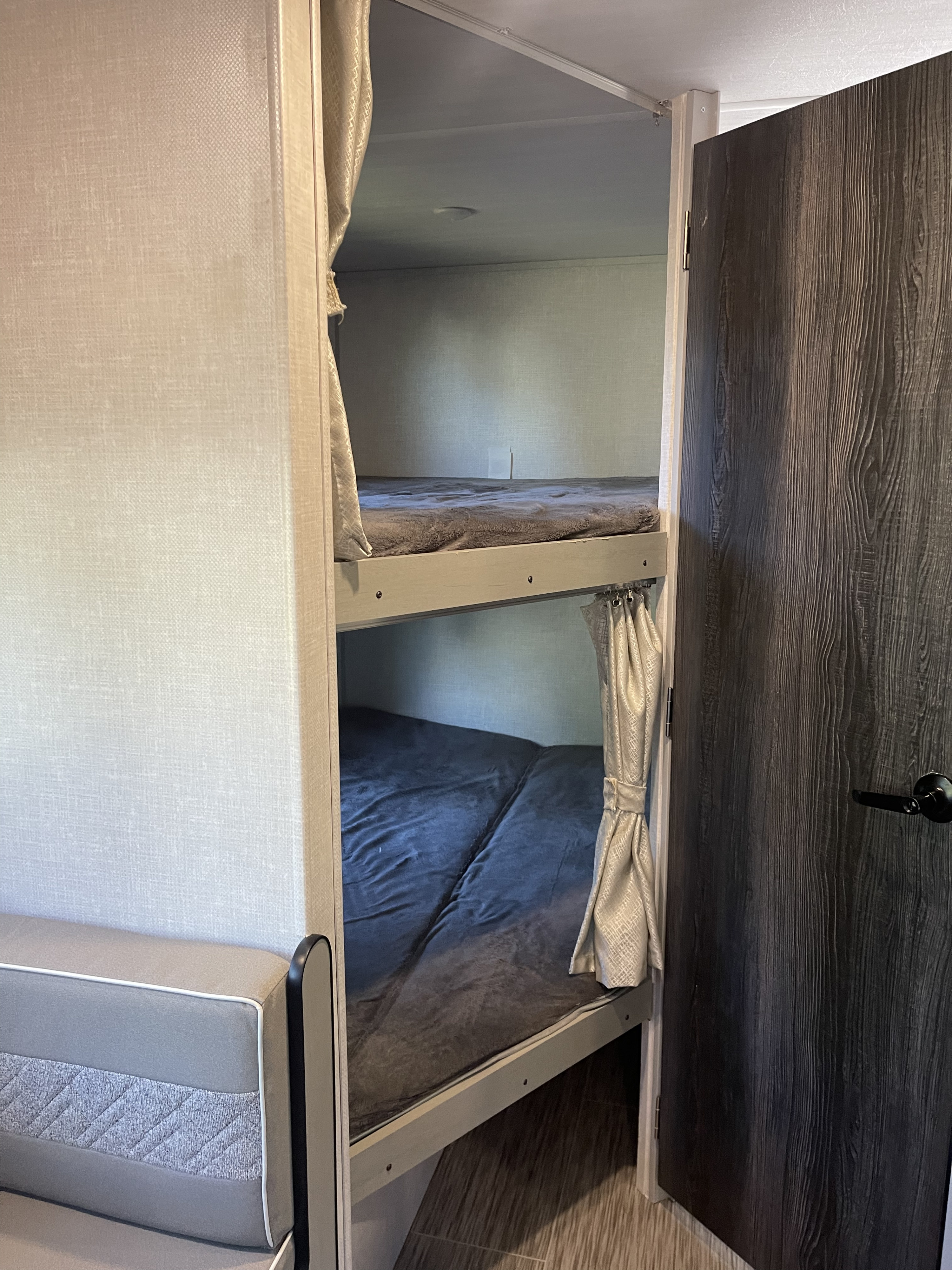 This RV rental interior showcases cozy bunk beds with privacy curtains, a grey seating area, and a dark wood-look door.. Forest River Ibex 2021
