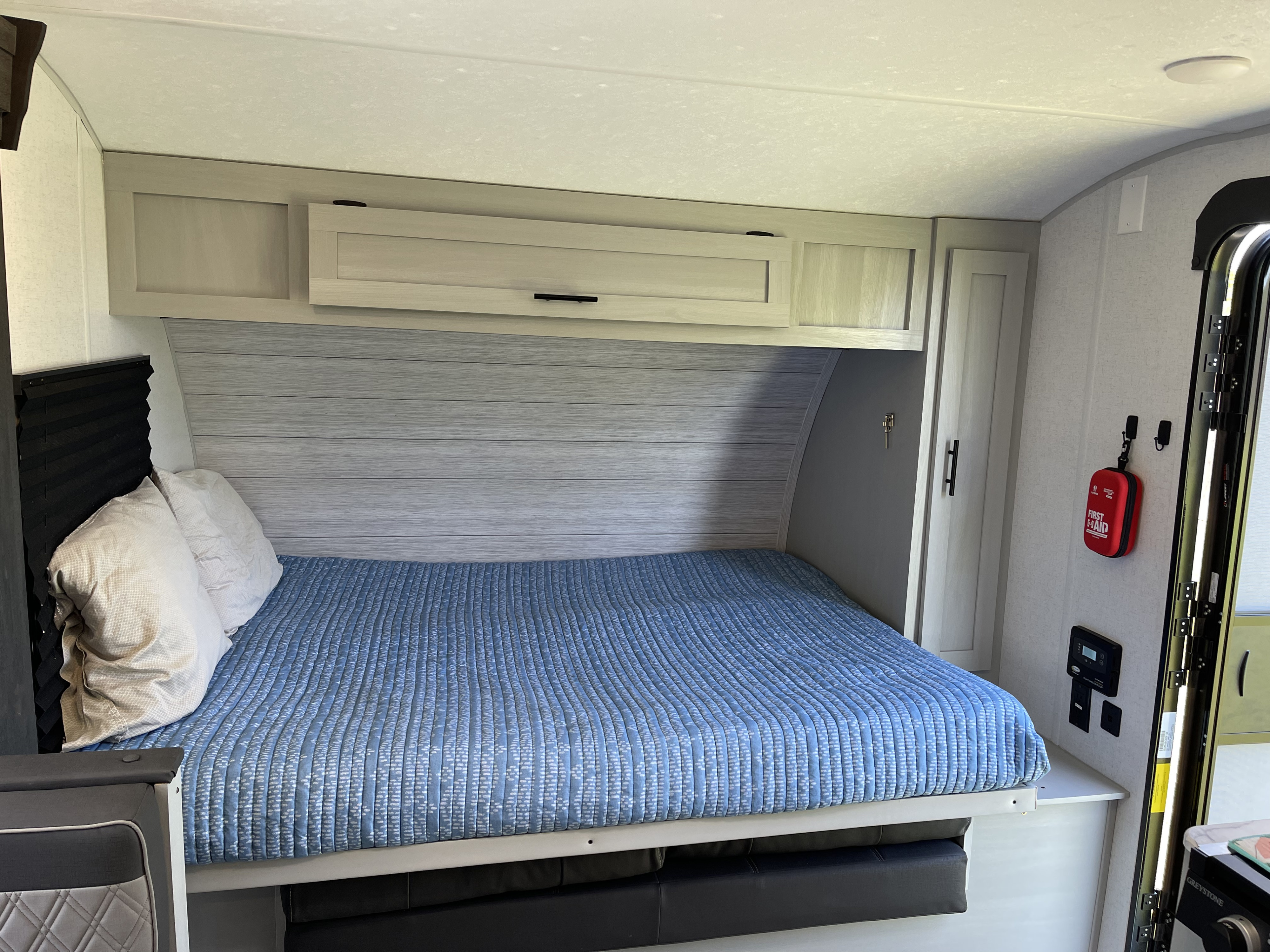 Interior of an rv rental features a cozy bed with a blue quilt, gray headboard, light overhead cabinets, and a red first aid kit.. Forest River Salem FSX 2022