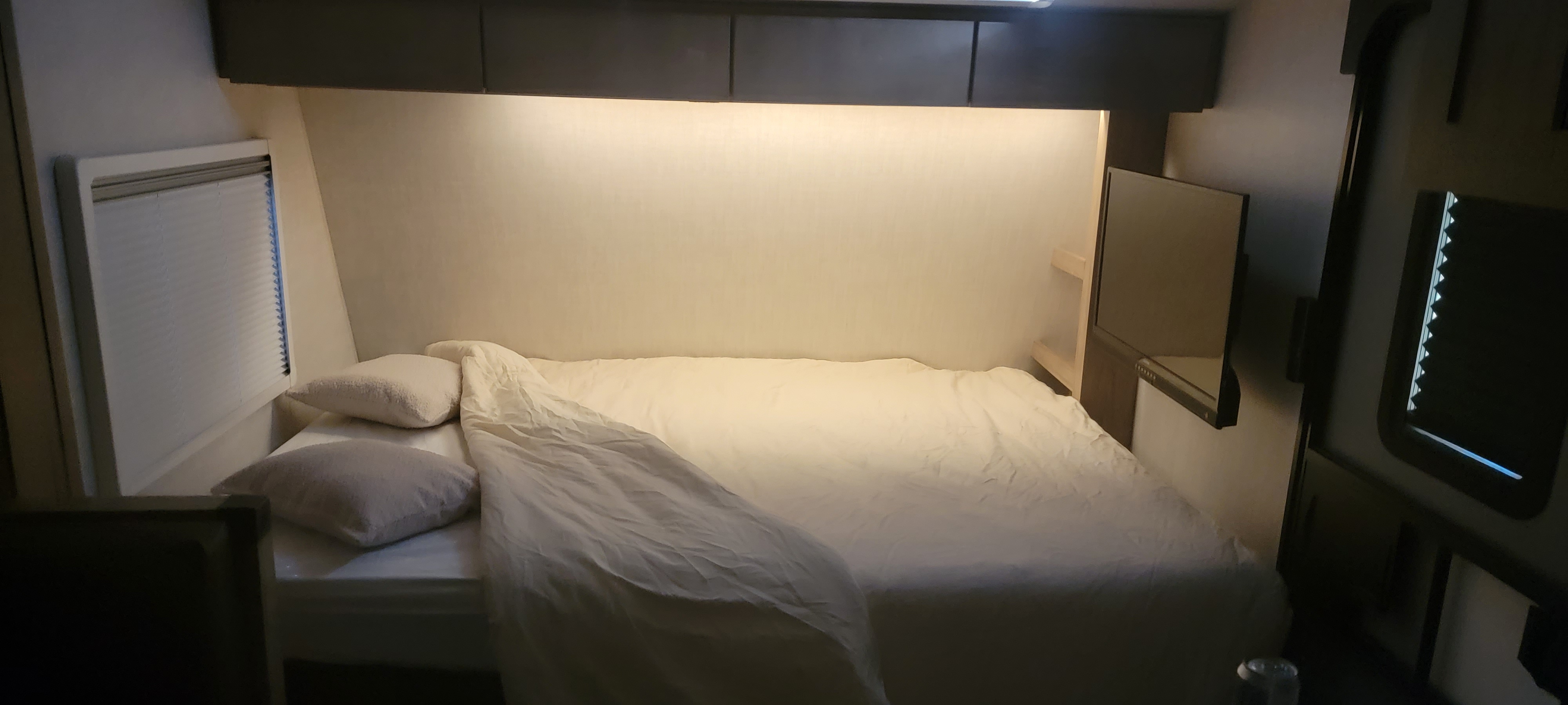 Inside an RV rental, a comfy bedroom with a neatly made bed, under-cabinet lighting, window blinds, and TV setup.. Ember Overland 2023
