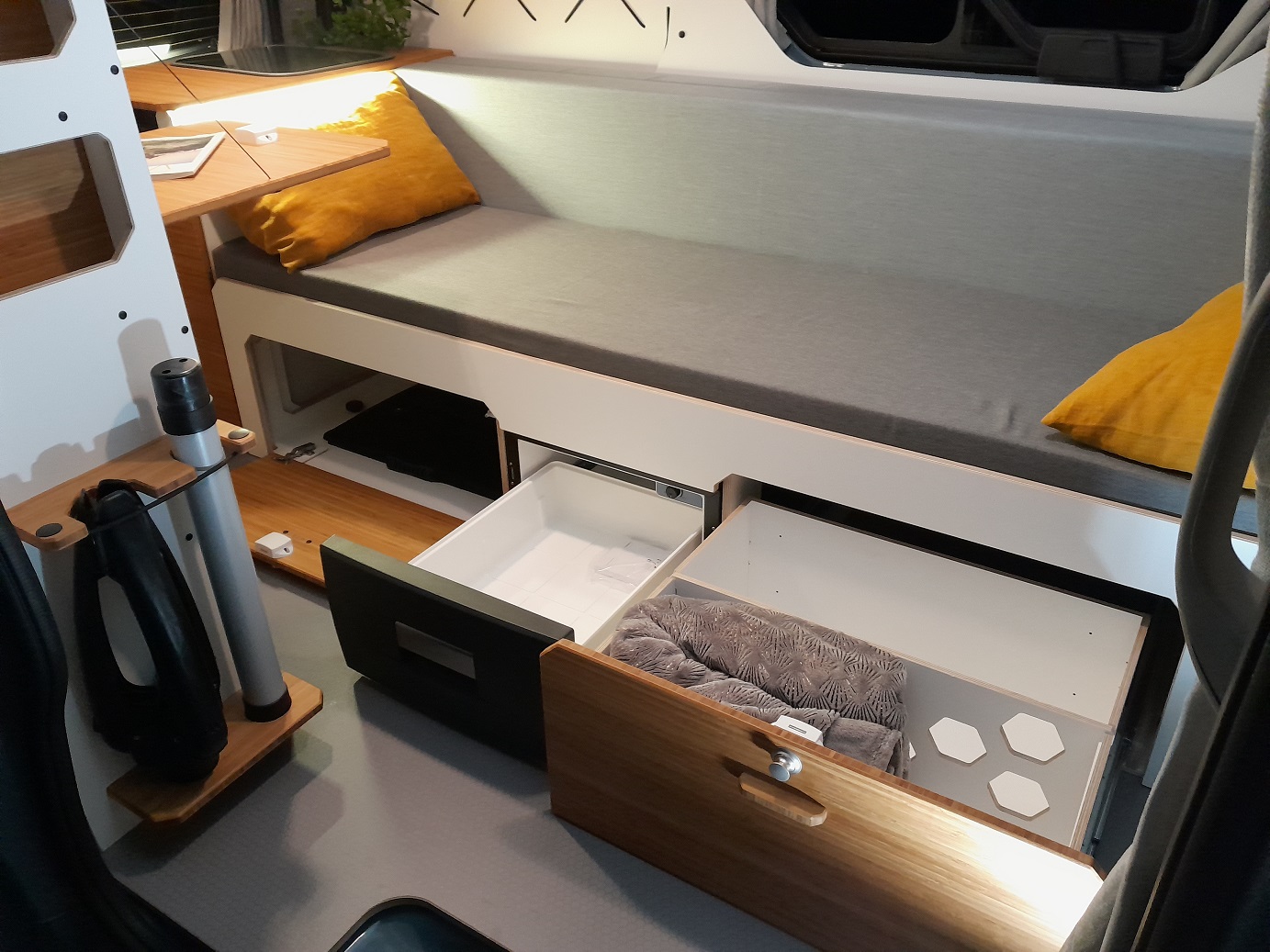 RV rental interior: A gray cushioned bed/bench with open storage drawers below, revealing a compact fridge and space for a blanket.. Renault Trafic aménagé VAN-MANIA North West 2023