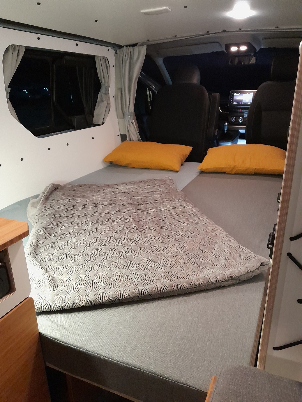 This RV rental offers a cozy camper bed setup with yellow pillows, grey blanket, and night views from the windows.. Renault Trafic aménagé VAN-MANIA North West 2023