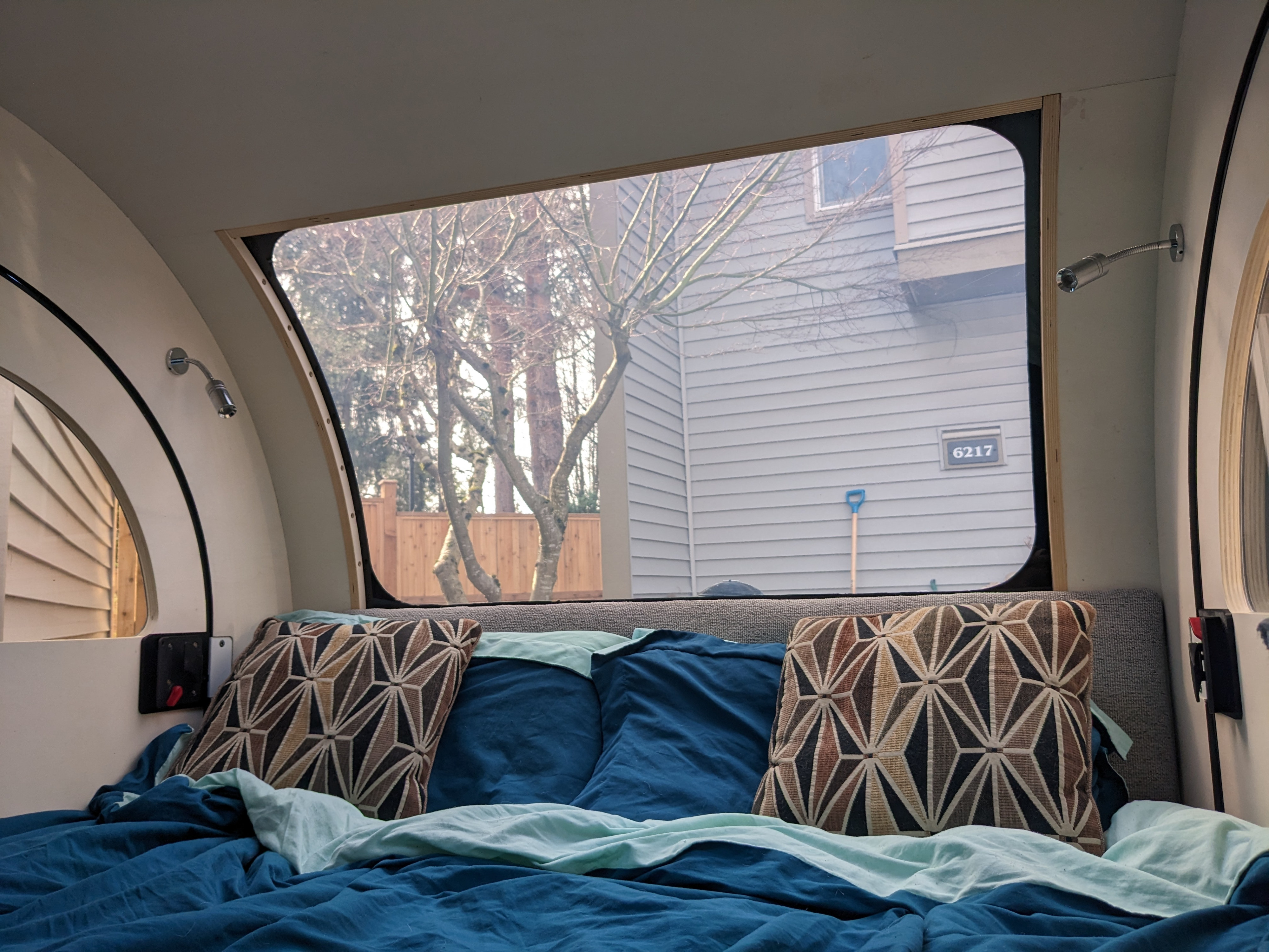 RV rental interior featuring a cozy bed, geometric pillows, and a large window showing bare trees and a nearby house.. Droplet Travel Trailer 2022