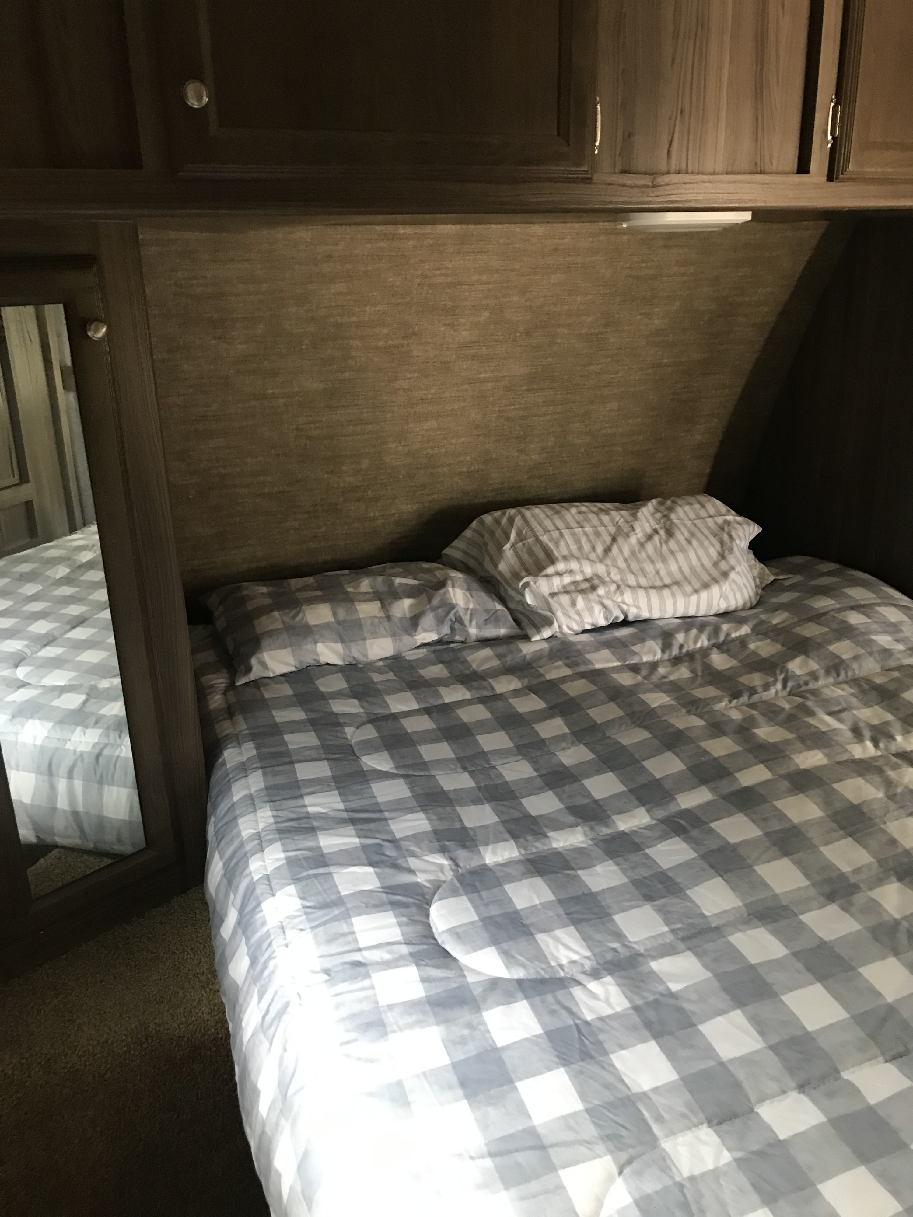 Inside an RV rental, a compact bedroom features a bed with checkered comforter, wood walls, overhead cabinets, and a mirror.. Coachmen Apex 2018