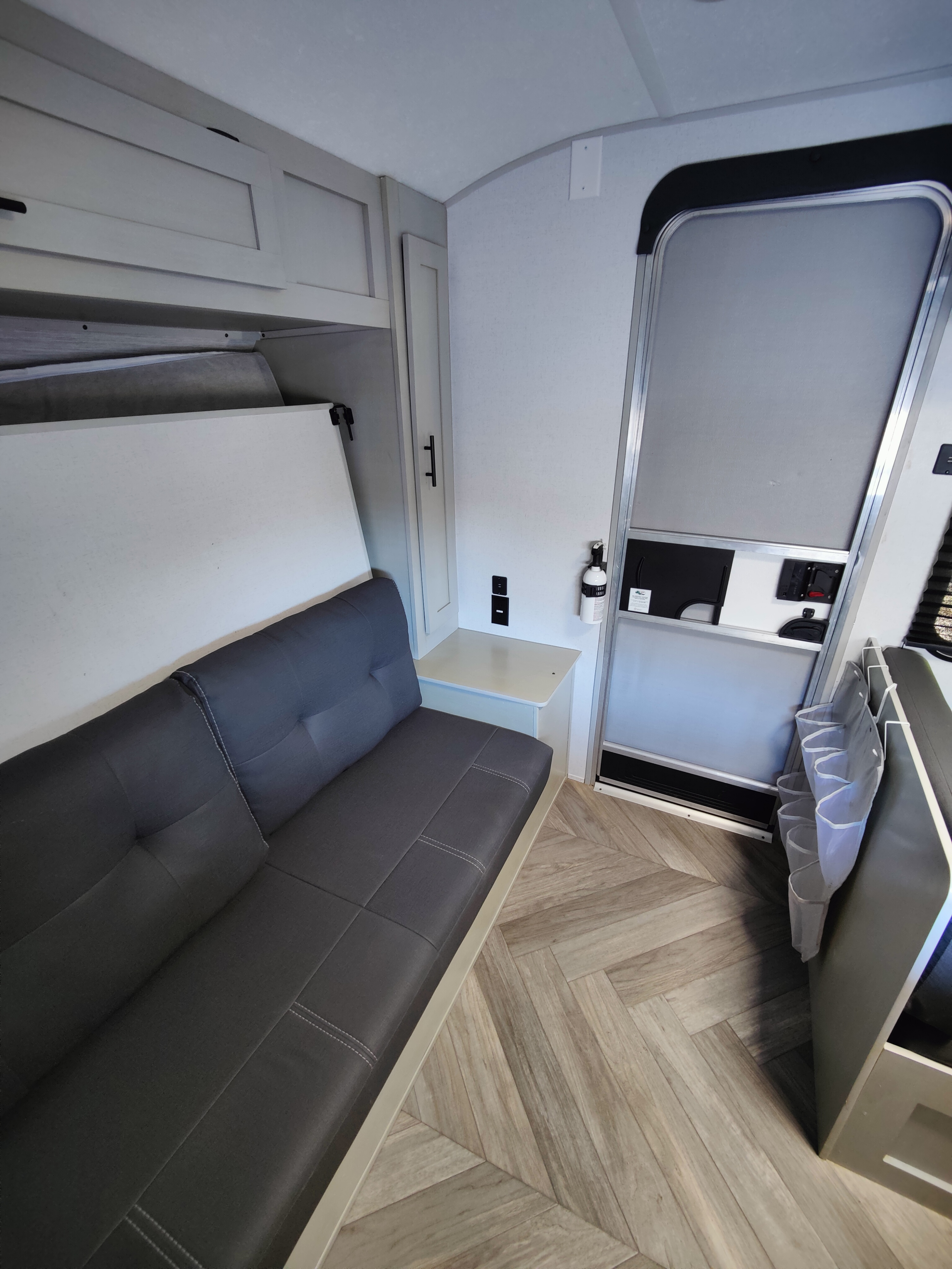 This rv rental showcases a gray convertible sofa, overhead cabinets, and herringbone flooring. Entry door and fire extinguisher too.. Forest River Wildwood FSX 2021