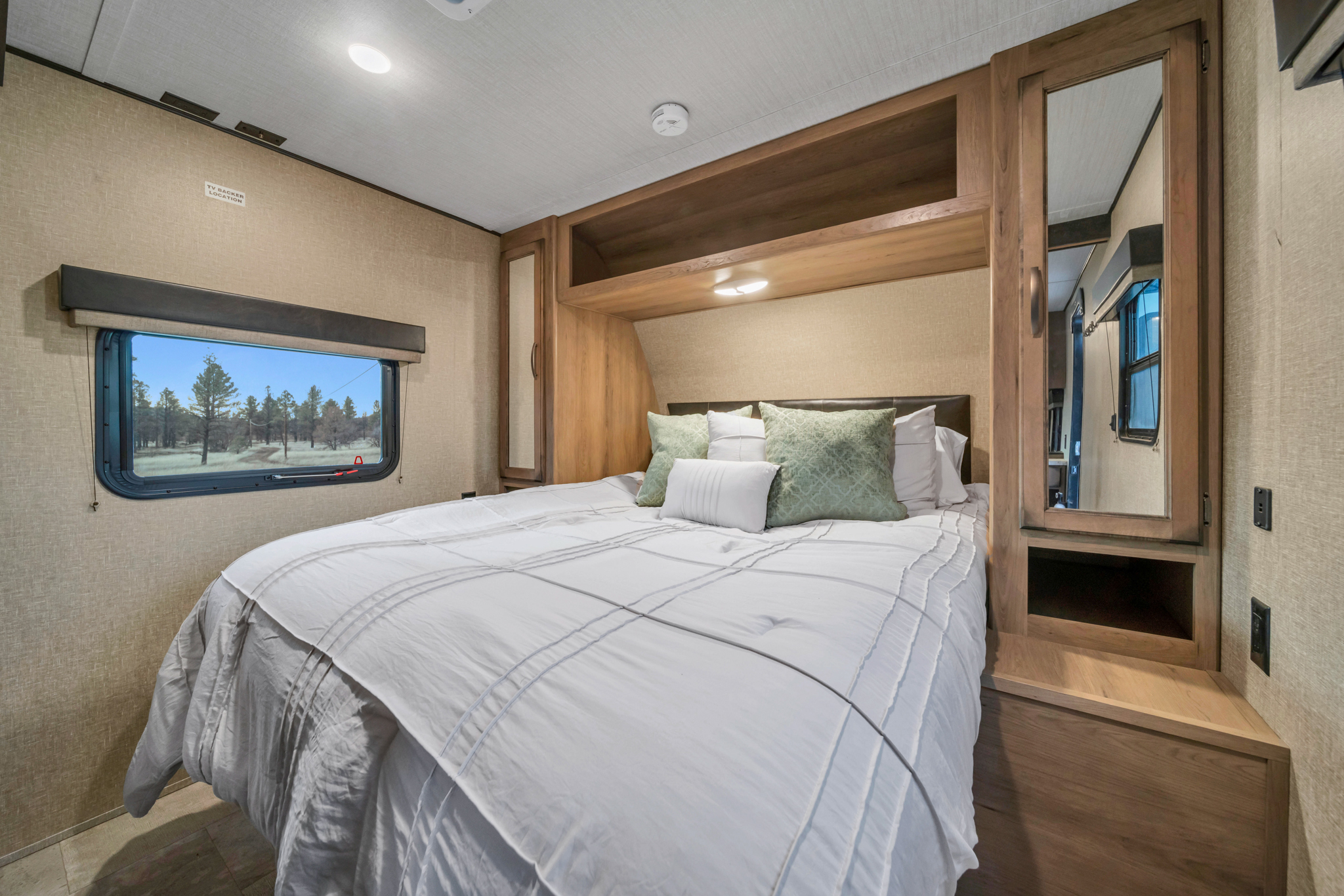 Modern RV rental bedroom: queen bed with green pillows, mirrored wardrobe, and window view of trees. Ideal for travel.. Grand Design Transcend Xplor 2020