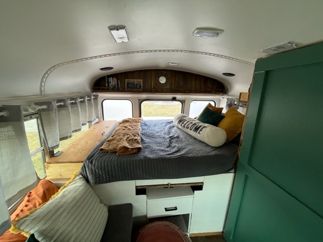 Cozy bedroom interior of a converted bus, ideal for your next RV rental. Features a raised bed, windows, and built-in storage.. GMC Savana 2002
