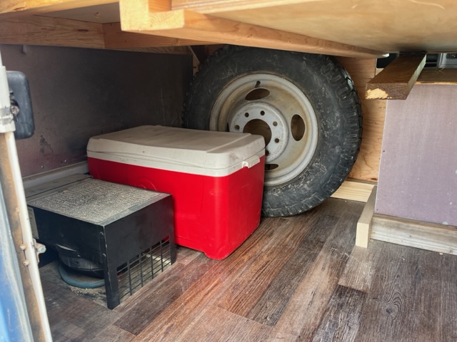 Under-bed storage in an RV rental: a spare tire, red cooler, and black heater on wood flooring.. GMC Savana 2002