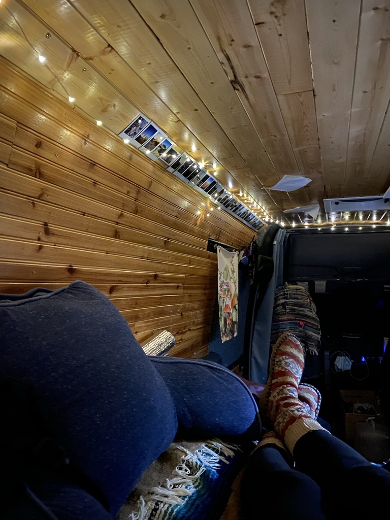Cozy RV rental interior with warm wood panels, string lights, and photos. Person relaxing, enjoying van life comfort.. Dodge Sprinter 2005